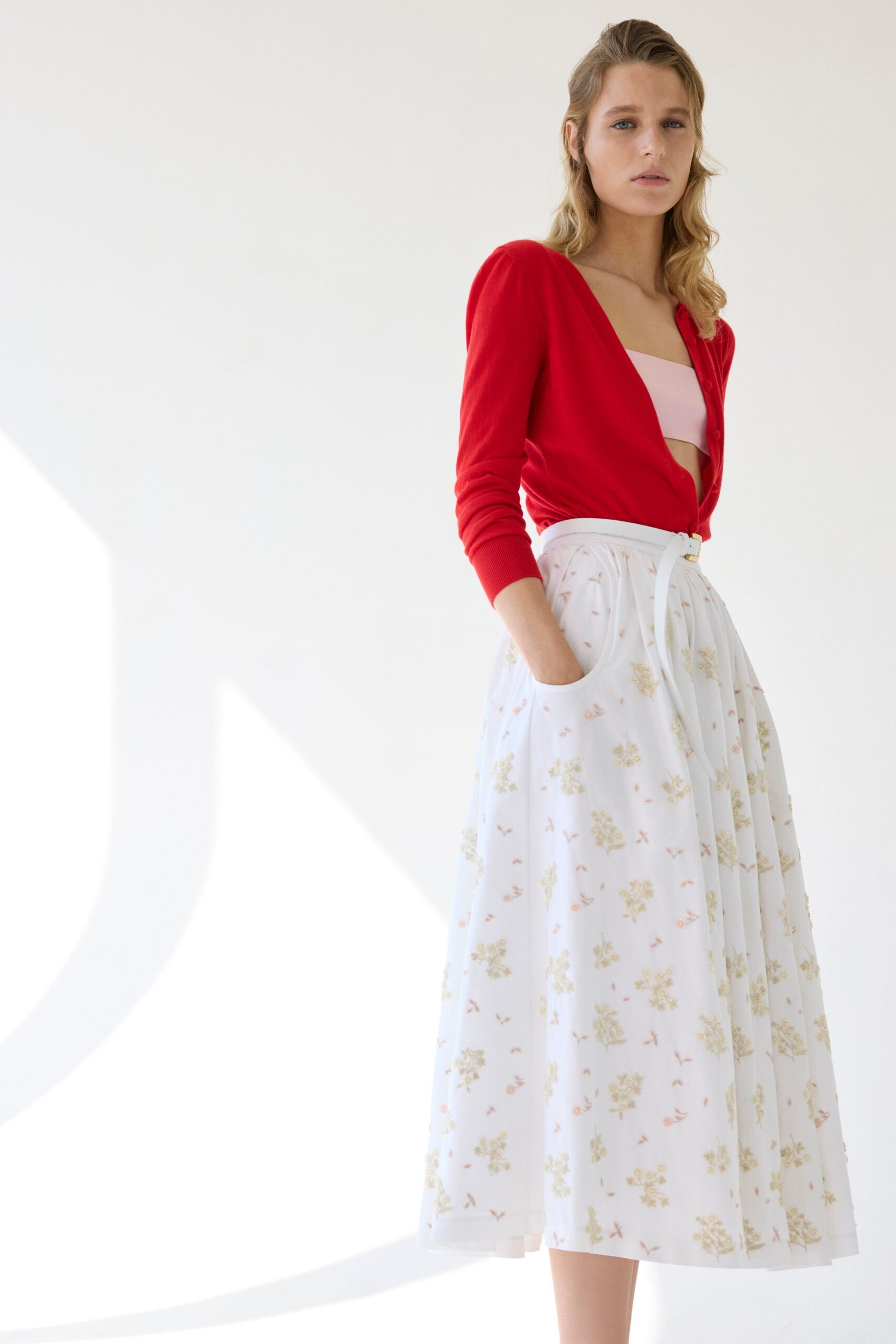 Cherry knit cardigan with rounded shoulders and tiny tonal buttons. Pale pink bandeau cutting a clean line across the chest. Ivory sheer midi skirt, softly gathered, scattered with faded floral sprigs. White belt with a small square buckle at the waist. Morning wedding registry run, florist water still drying on the sidewalk.