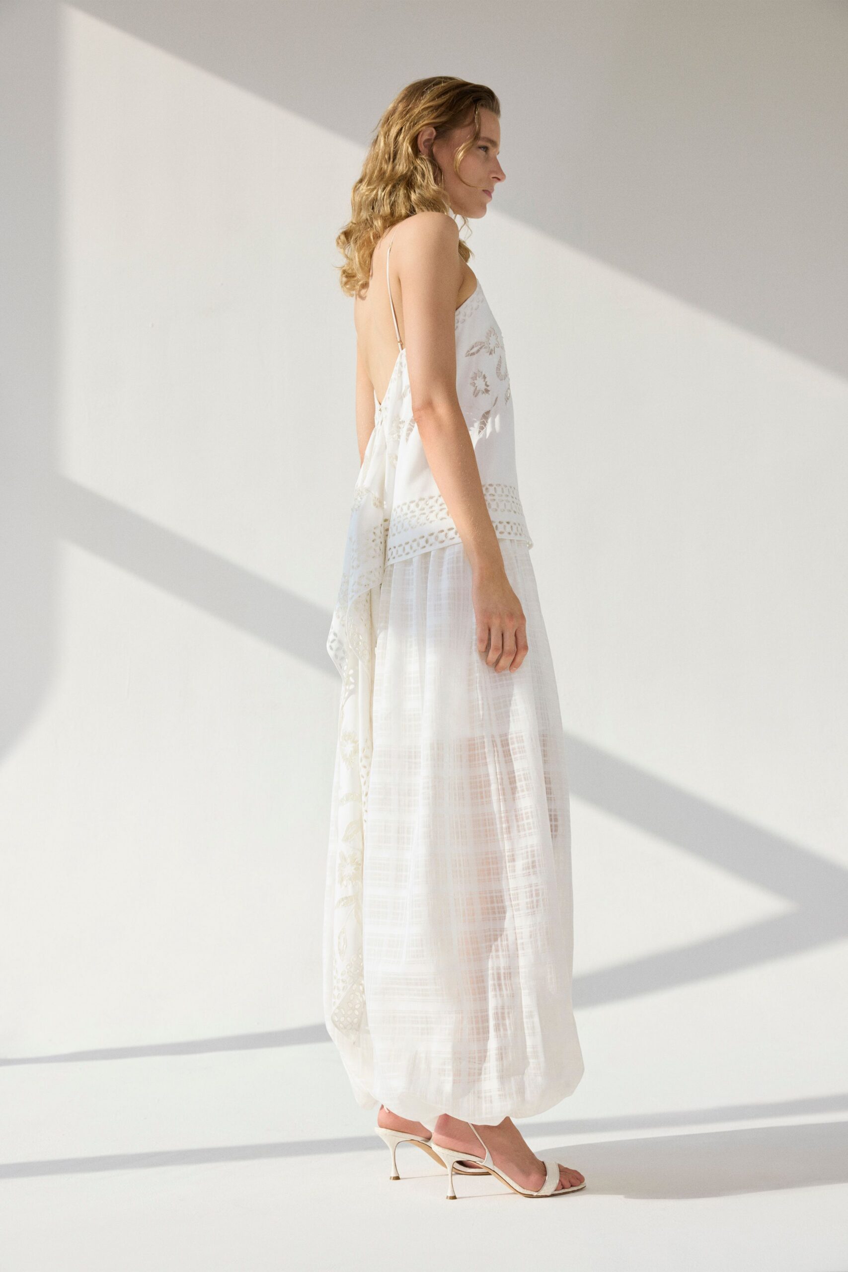 Ivory camisole with needlework flowers, ladder-cut hem and a low bare back. Sheer ivory gauze skirt, ankle length with a faint crosshatch weave. White thong mules on narrow flared heels. Morning in a rental house, sun stripes across the wall before anyone else wakes.