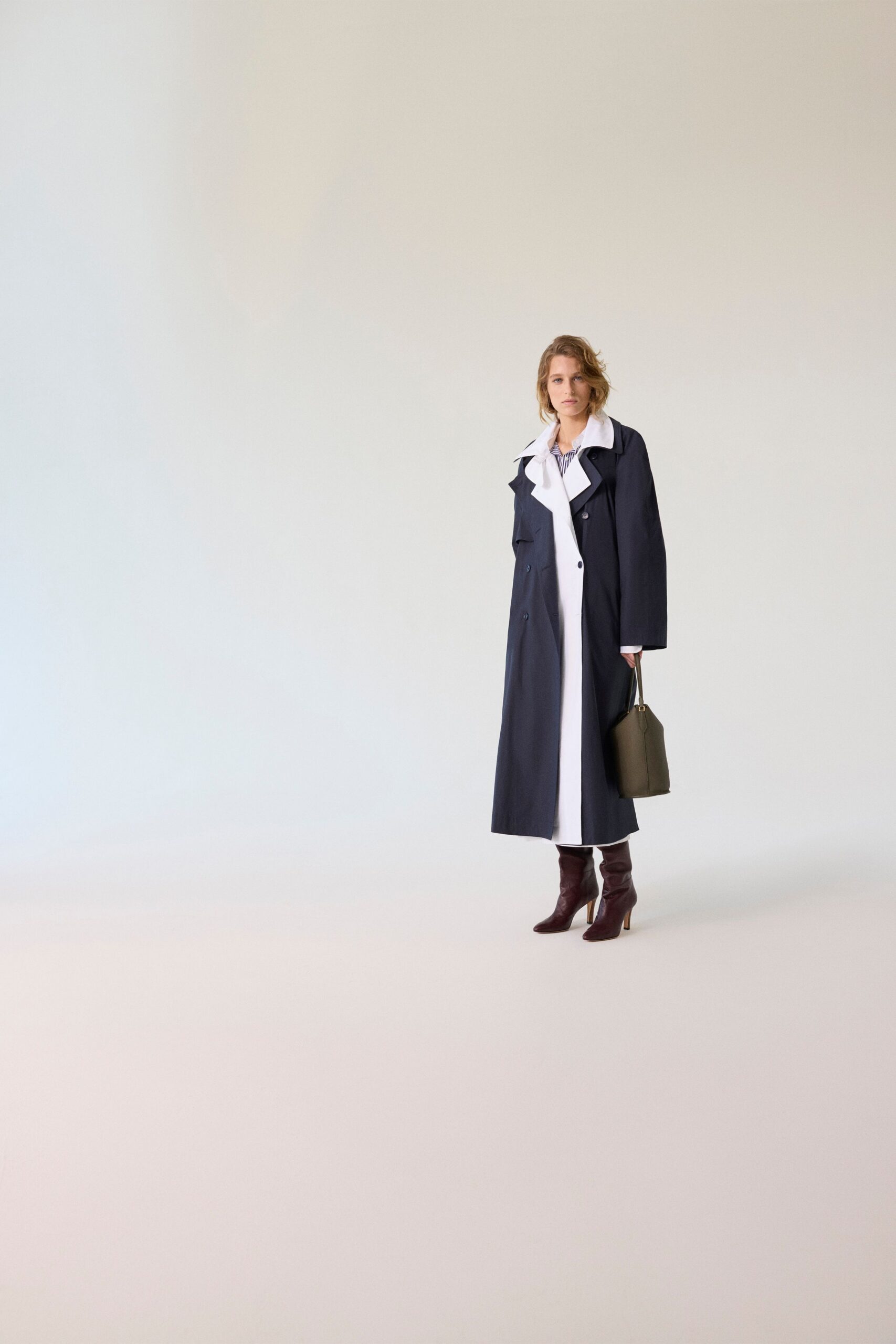Navy trench falling to the calf, white underlayer peeking through the front. Blue-and-white striped shirt, collar spread wide over the lapels. Olive trapeze tote with slim handles and stiff sides. Burgundy leather boots, gently slouched at the ankle. Walking into a courthouse early, coffee cooling in a paper cup.
