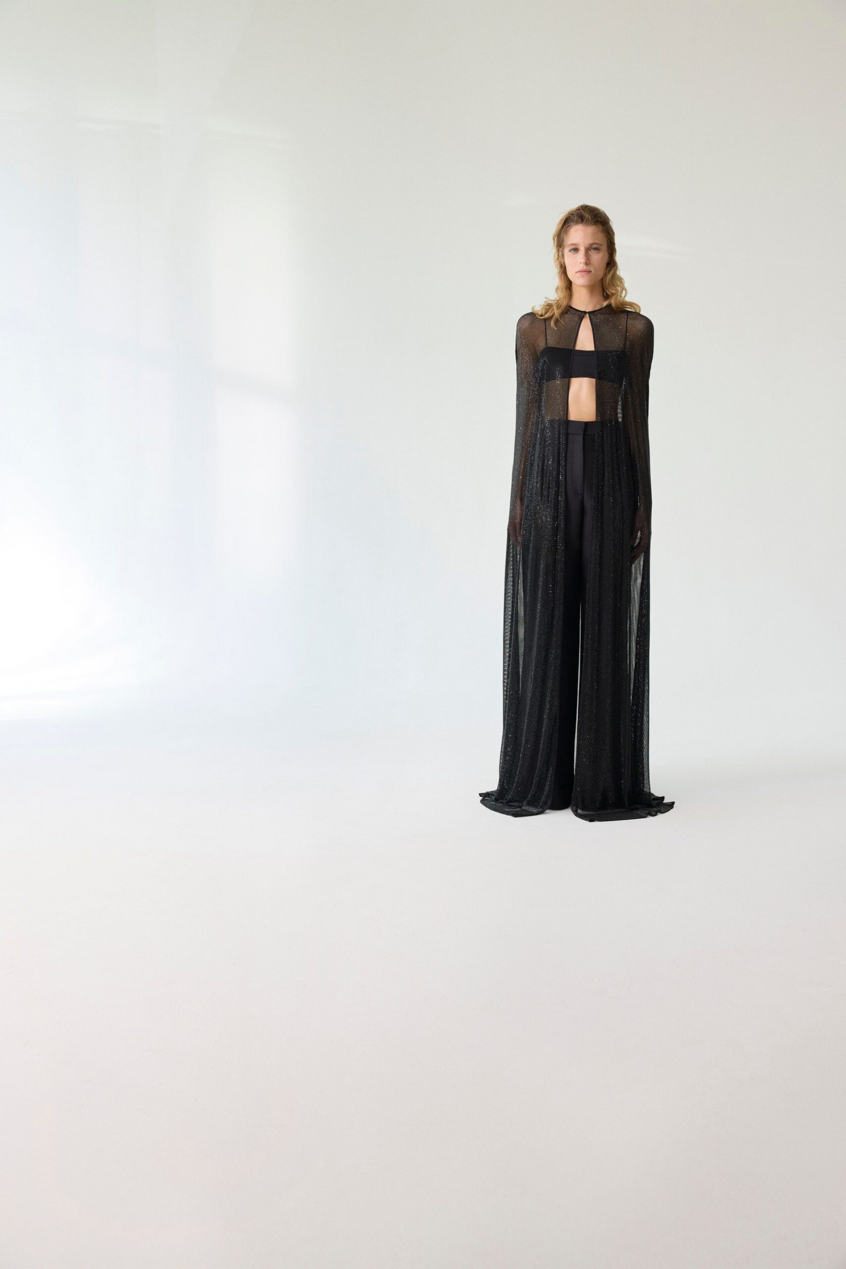 Black glitter mesh falling from a narrow keyhole neckline to the floor. Matte black bandeau cutting a blunt line across the ribs. High-waisted black trousers, pressed straight under the sheer overlay. Waiting barefoot by hotel curtains after the party finally thinned out.