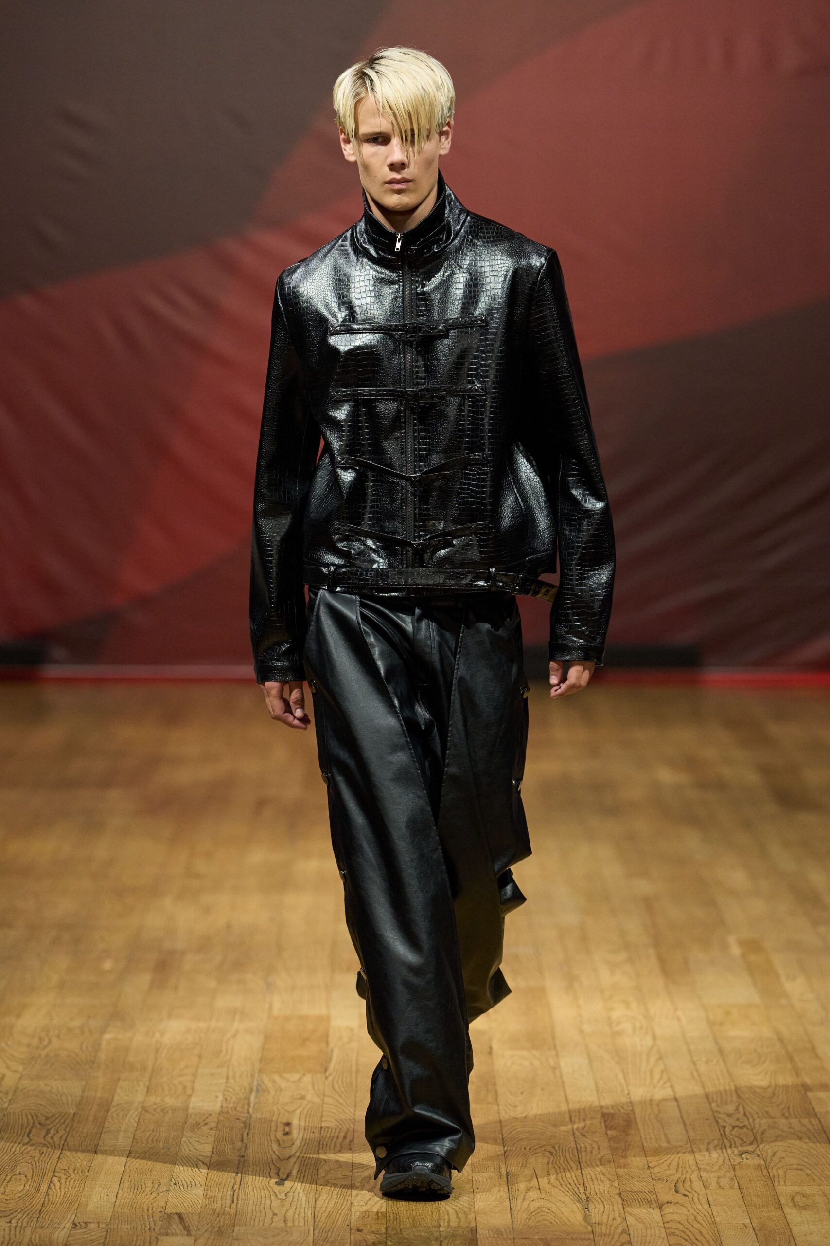 Black crocodile-embossed leather jacket with frog closures crossing the zip. Wide black leather trousers, snap tabs climbing the outer seams. Square black shoes, barely visible under the pooled hem. Walking across a varnished hall floor after midnight rain.