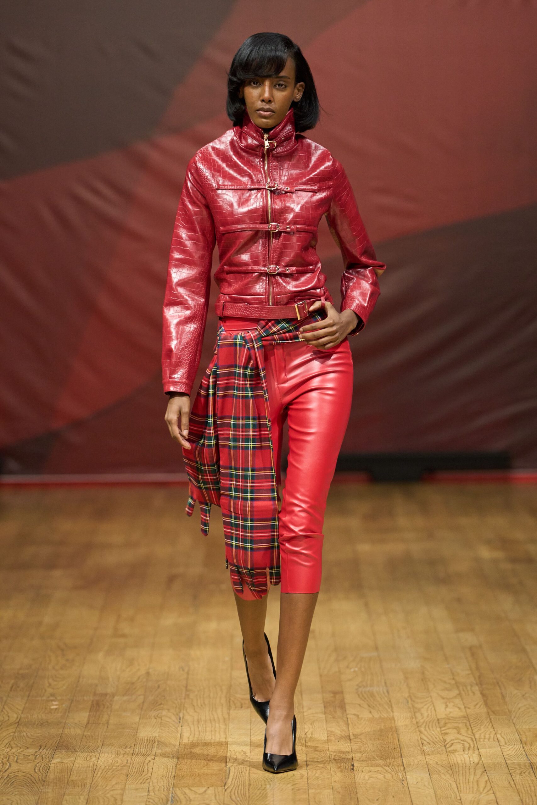 Glossy cherry croc-embossed leather jacket, zipped high with three buckle straps. Red leather capris, cut close and cropped mid-calf. Red tartan shirt knotted low at the hip, one tail hanging longer. Black pointed pumps. Walking out of a pub quiz still wearing the tablecloth dare.