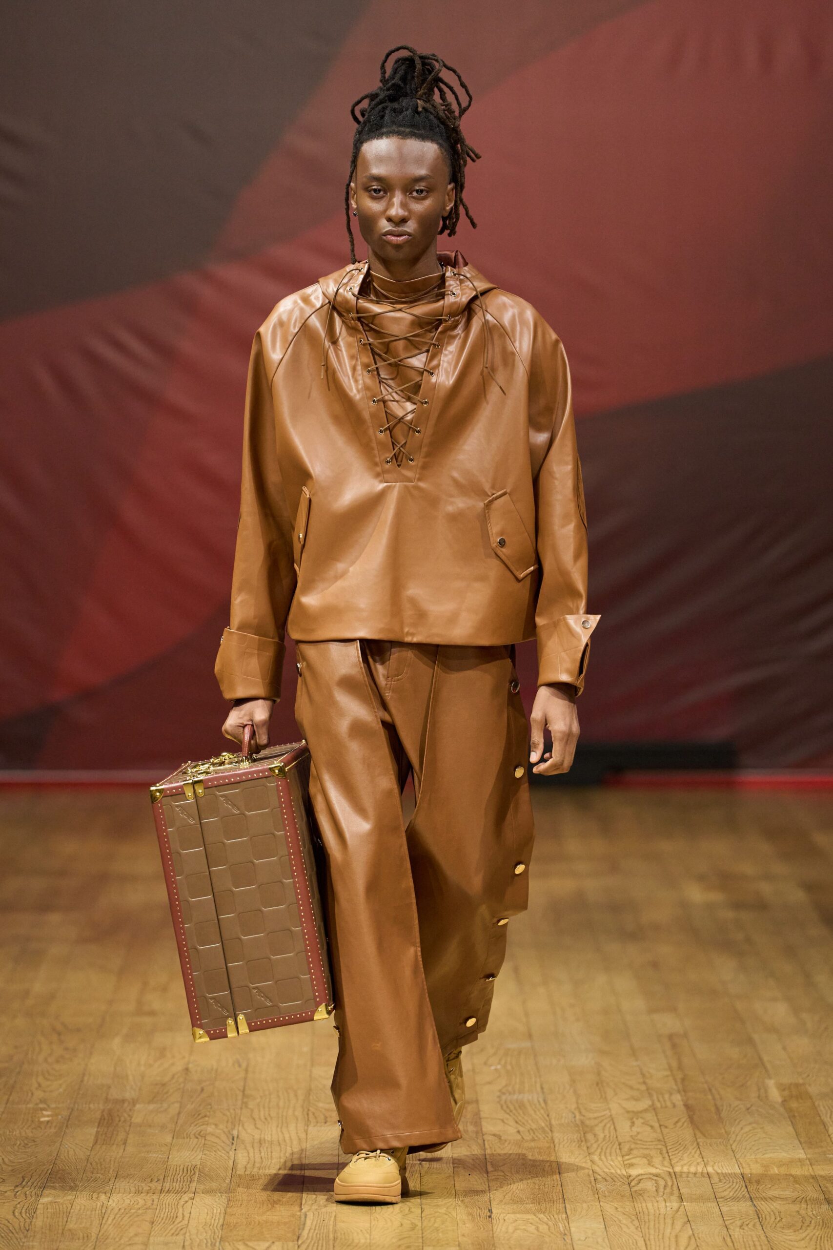 Cognac leather anorak with crossed lacing plunging below the throat. Matching wide leather trousers, brass snaps marching up the outer seam. Wheat work boots with thick padded collars. Hard suitcase embossed in a square grid with brass corners. For walking into a station hotel before dawn with nowhere smaller to pack.