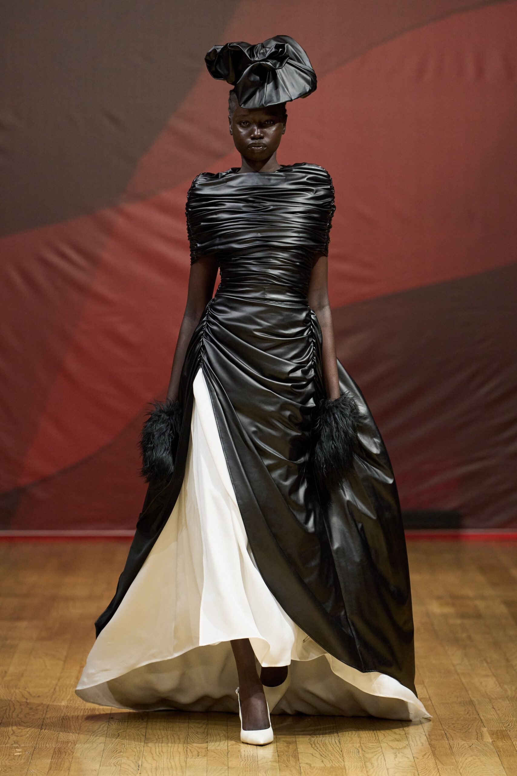 Oversized black satin bow hat, folded into stiff petal-like lobes. Black ruched satin gown with a wide white underlayer flashing at the front slit. Black feather cuffs at each wrist. White pumps, pointed and nearly swallowed by the hem. For descending a hotel staircase after midnight with everyone already staring.