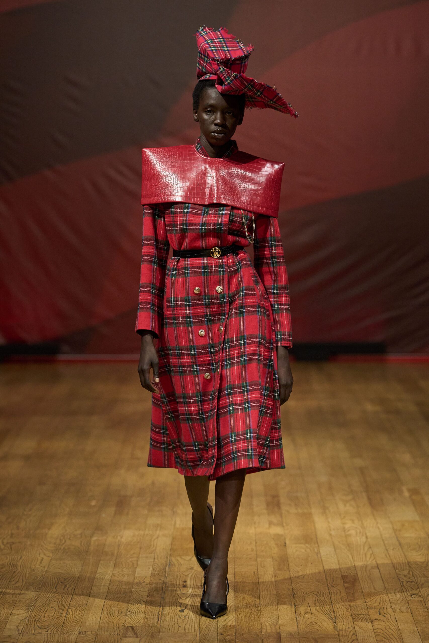 Oversized red tartan hat, one corner jutting out with frayed edges. Glossy red crocodile-embossed leather folded broad across the shoulders. Red tartan dress, bloused bodice above a buttoned kilted skirt. Black belt with a round gold medallion buckle. Black pointed pumps. Leaving a Burns supper after the speeches ran long.