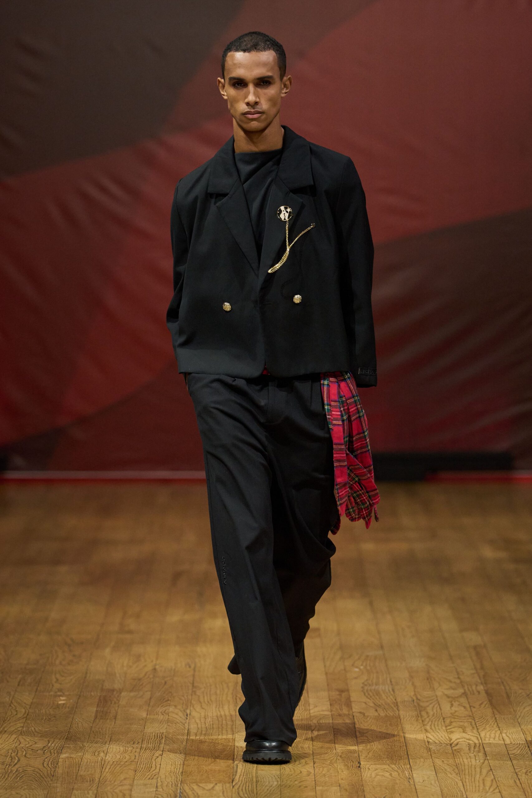 Black double-breasted blazer with gilt buttons and a draped chain brooch. Black top with a high folded neckline. Loose black trousers with a red tartan shirt tied at one hip. Square-toe black shoes with thick tread soles. Leaving the student union after midnight, still wearing someone's picnic blanket.