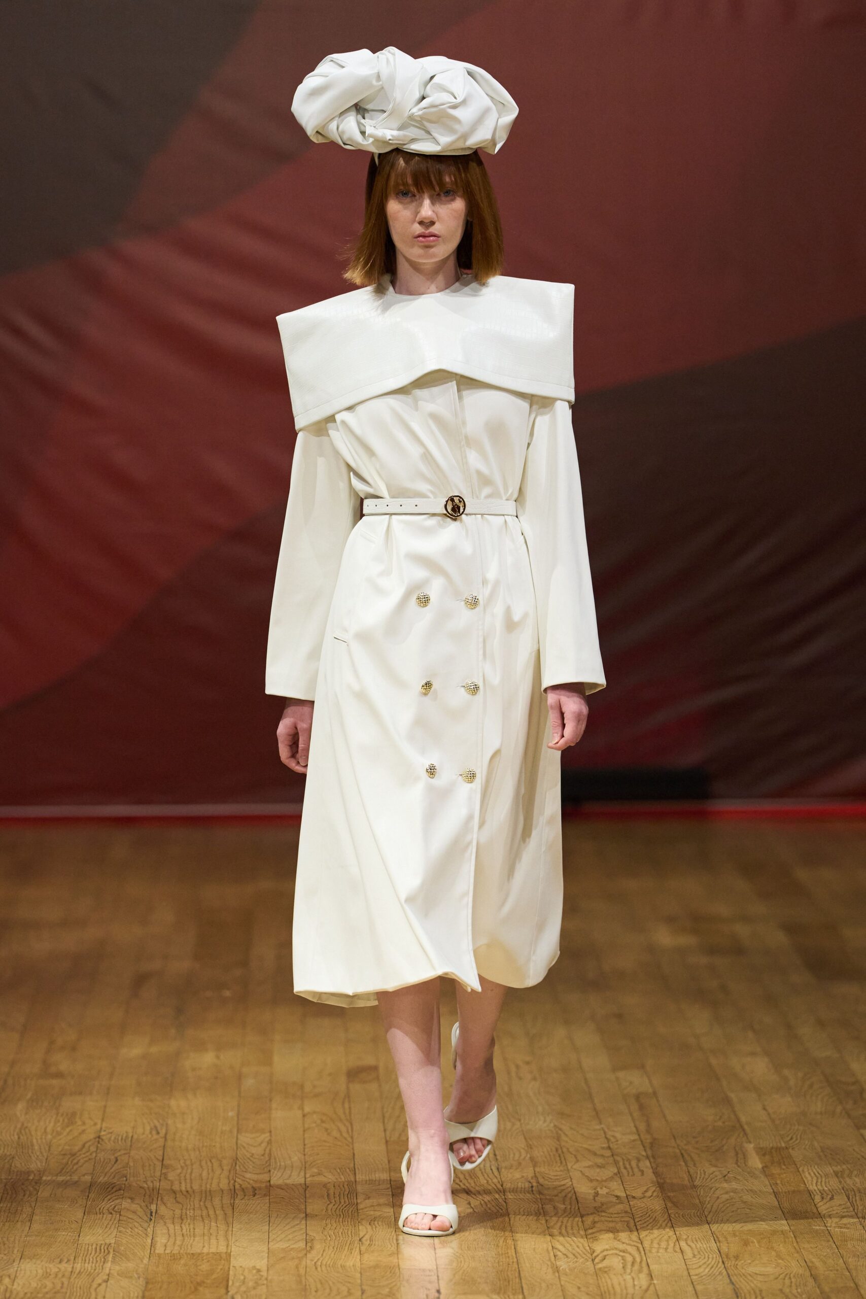 Oversized white turban knot in crisp folded cloth. Ivory satin coat dress with a squared cape yoke and six textured buttons. Narrow white belt with a round gold buckle at the waist. White open-toe mules with broad curved straps. Registry office at noon, bouquet paper still crackling in your hand.