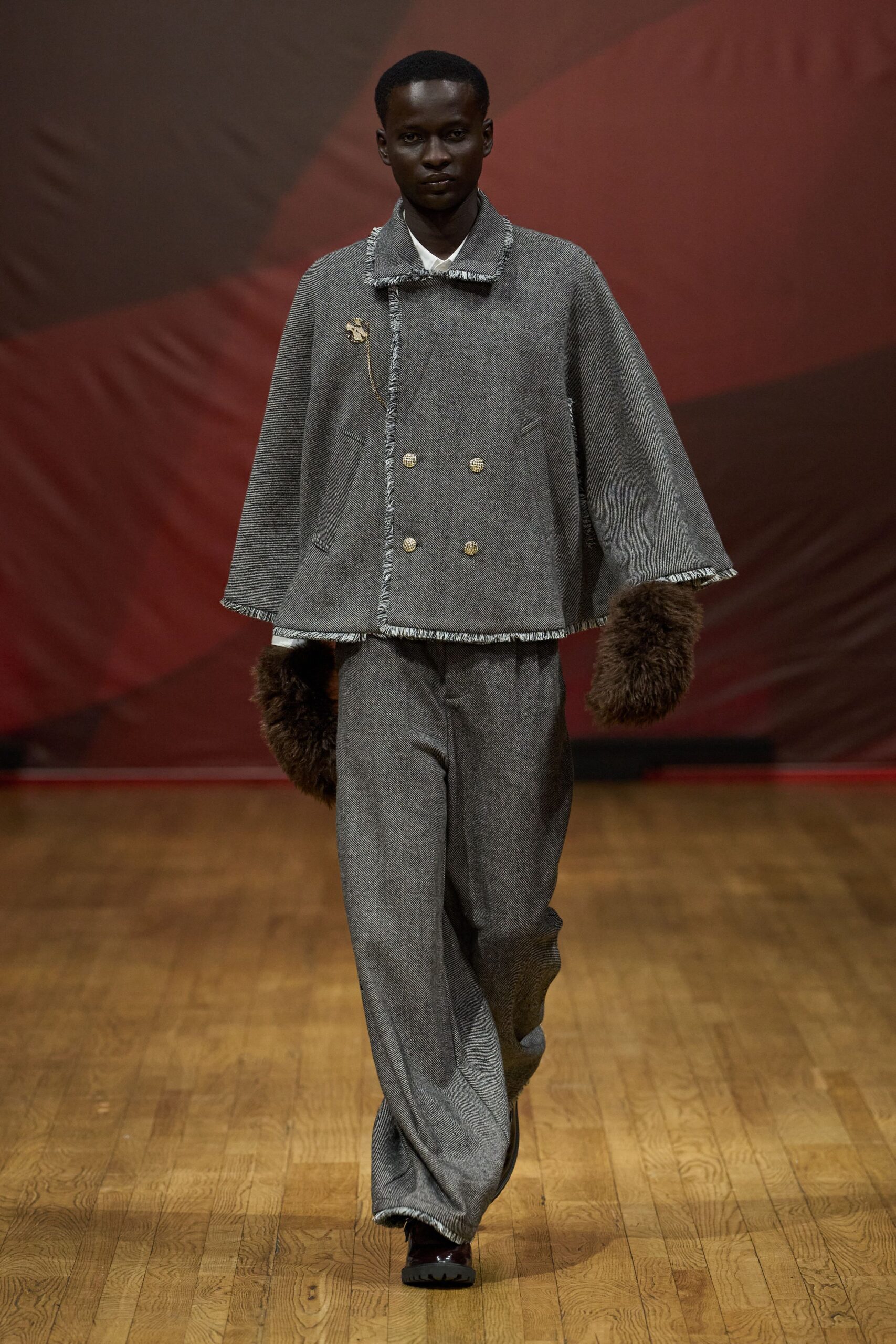 Grey herringbone cape jacket with frayed edges and four crest buttons. White shirt point peeking cleanly at the throat. Dark brown fur gauntlets bulk out both wrists. Loose grey herringbone trousers breaking heavily over the shoe. Burgundy leather shoes with a lug sole. Crossing a drafty lodge hall before dinner is called.
