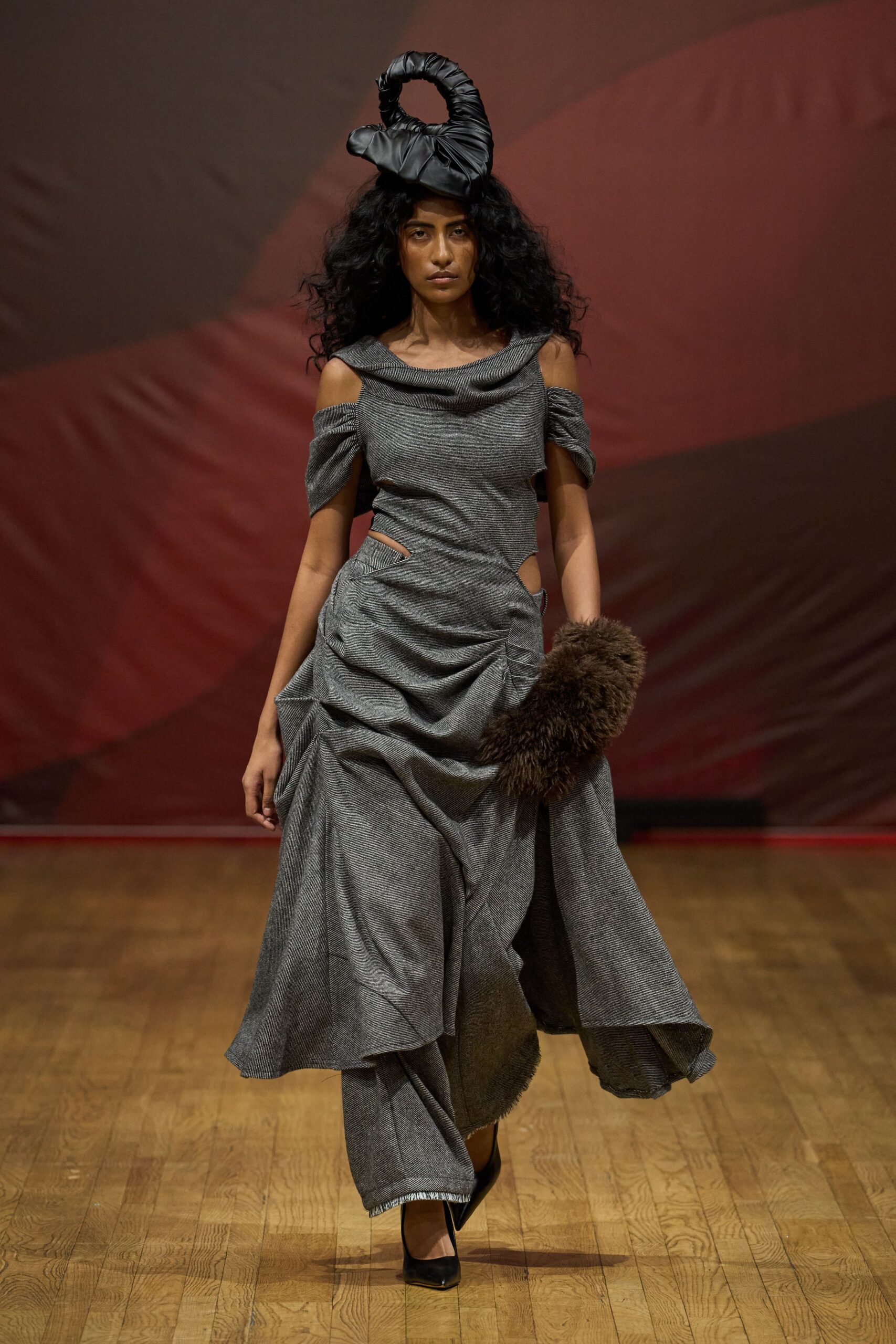 Black padded spiral looped upright like a twisted inner tube. Grey micro-herringbone dress with a folded cowl, hip cutouts and ruched drape. Dark brown fur muff covering one hand. Black pointed pumps. Crossing a draughty civic hall with the raffle already underway.