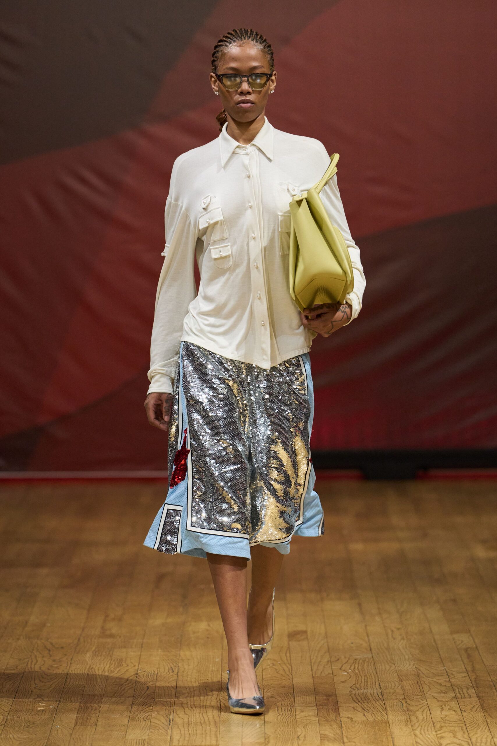 Olive-tinted rectangular lenses. Ivory jersey blouse with pearl buttons and flap chest pockets. Silver sequin panel skirt edged in powder-blue mesh, red tabs flicking out. Butter-yellow folded leather tote tucked under the arm. Mirror-silver pointed pumps. Walking into a hotel lobby with flowers and one garment bag left.