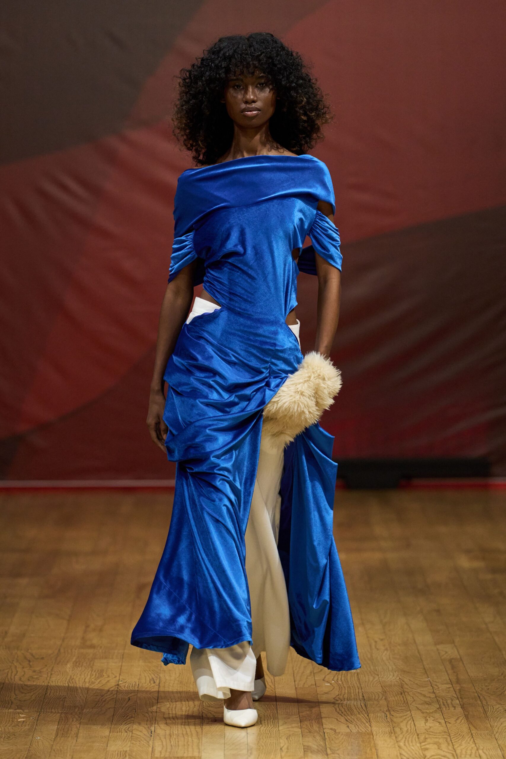 Electric-blue satin gown with a folded off-shoulder band and ruched cutout waist. Cream wide-leg trousers breaking below the gown's high front split. Shaggy cream fur muff tucked at the hip. White pointed heels peeking out in short steps. Crossing a hotel ballroom after midnight with your gloves still off.