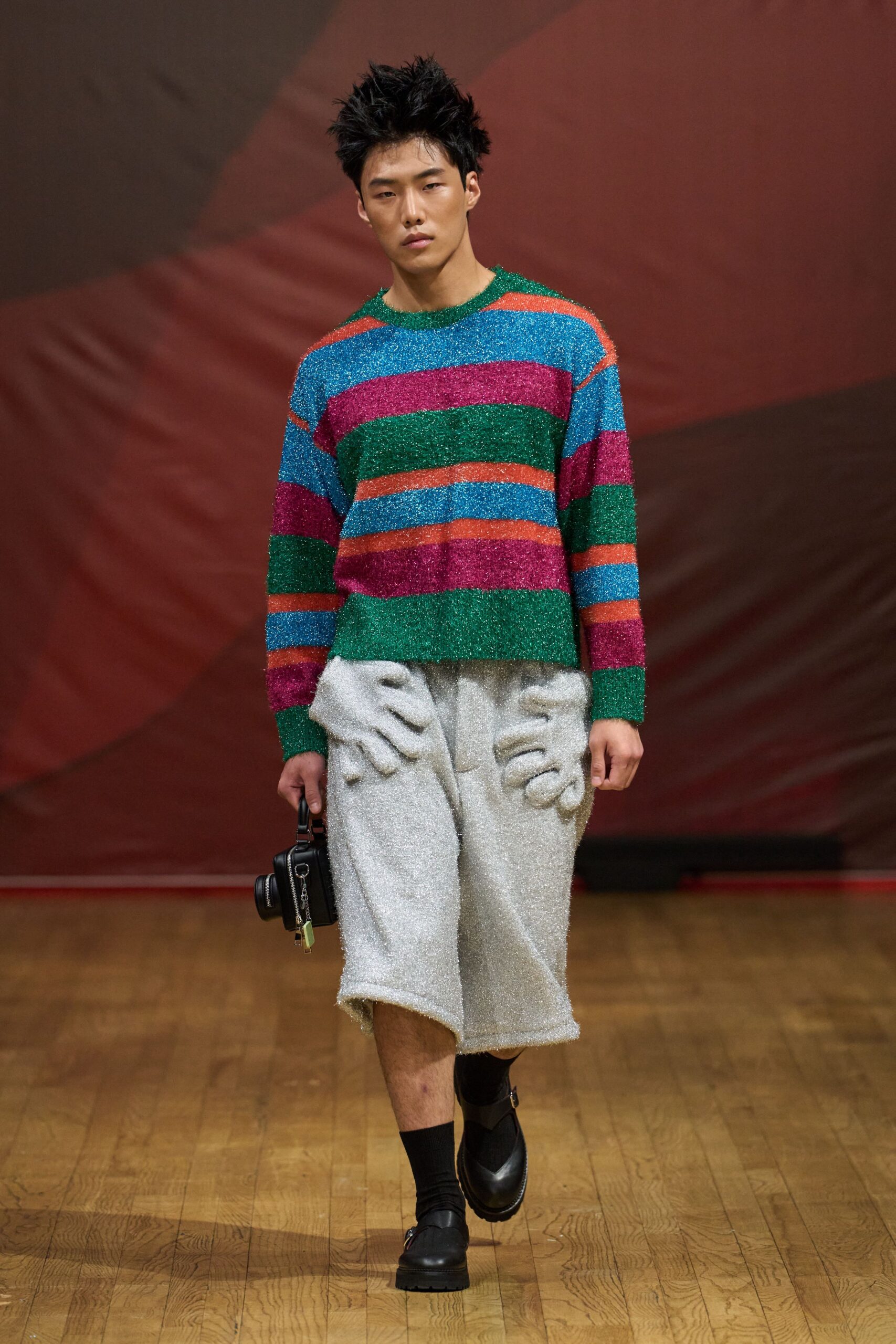 Striped lurex sweater in cyan, emerald and fuchsia, collar softly fuzzed. Silver bouclé knee shorts with padded hand shapes swelling over the hips. Boxy black camera bag with a dangling green tag. Black rib socks. Black monk-strap shoes with heavy tread soles. Walking into a holiday party still carrying the office camera.