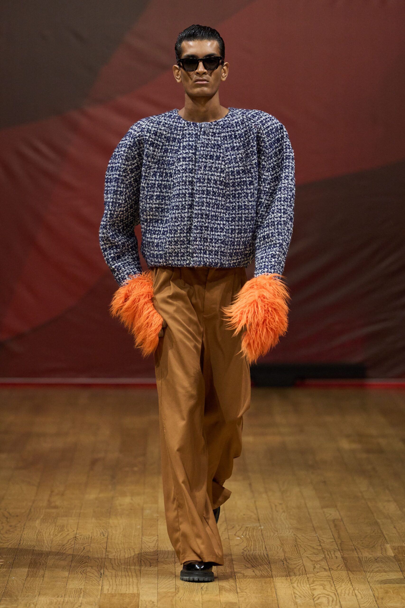 Black rectangular sunglasses. Blue-and-white bouclé pullover with a wide boat neck. Electric-orange shaggy fur cuffs swallowing the hands. Caramel wide-leg trousers with a sharp front crease. Black lug-sole shoes peeking from the hem. Walking into a hotel lobby still wearing feathers from the car seat.