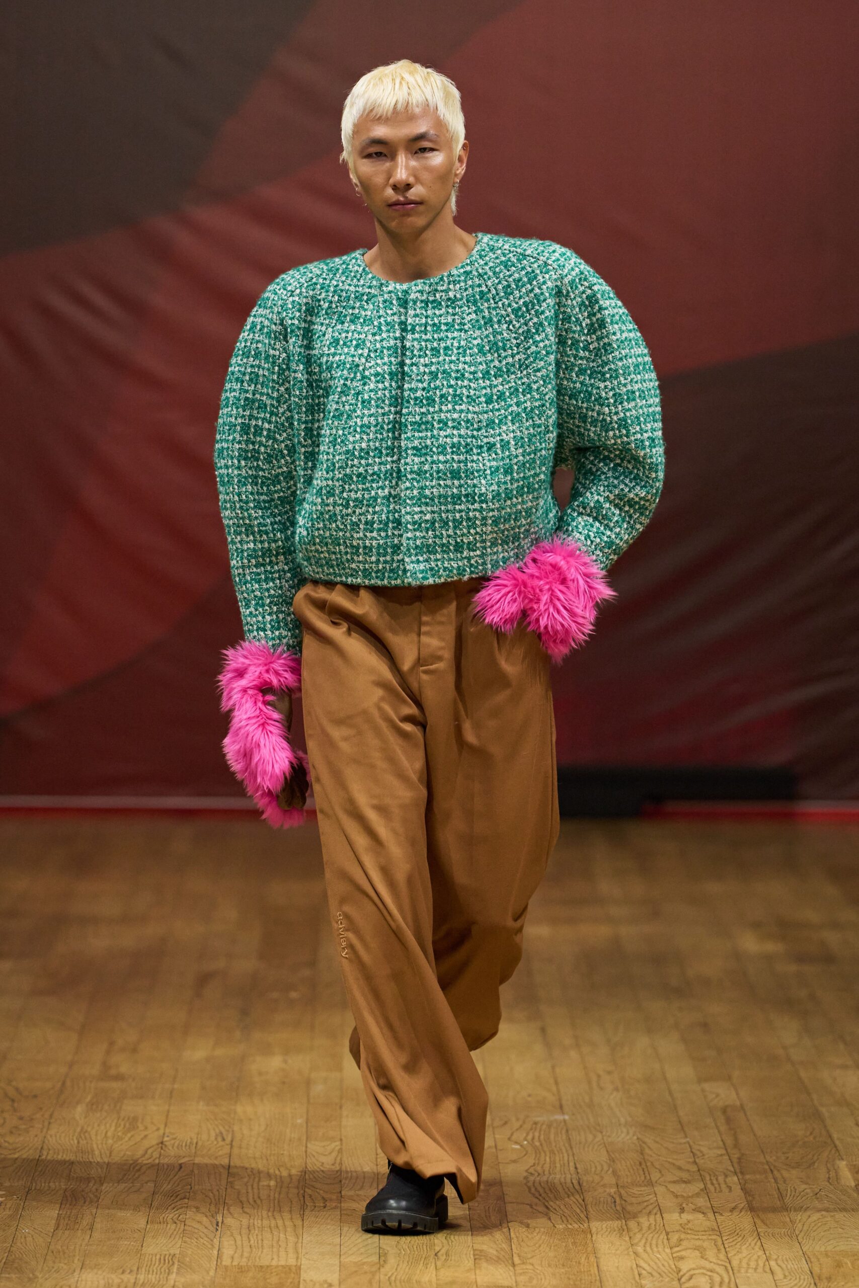 Boxy jade-and-cream bouclé top with padded shoulders and hidden front closure. Hot-pink shaggy faux-fur gauntlets swallowing both wrists. Caramel trousers cut full through the leg with a soft front pleat. Black square-toe shoes with a chunky lug sole. Walking into a hotel bar still carrying static from a borrowed mohair blanket.