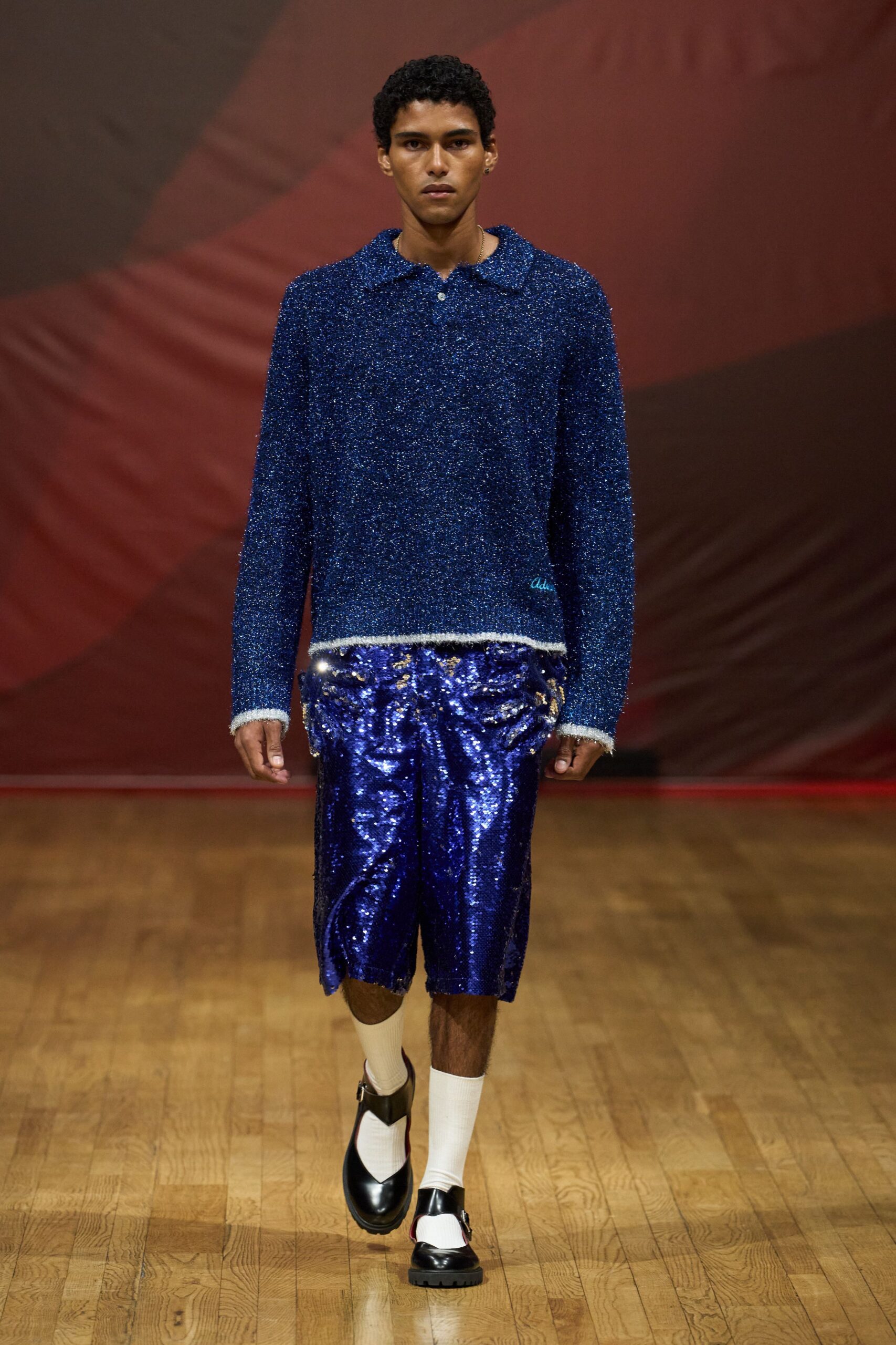 Cobalt lurex knit polo, fuzzy white trim at cuffs and hem. Knee-length paillette shorts in electric blue, crumpling like foil. Cream ribbed mid-calf socks. Black Mary Jane loafers with a white vamp panel. Walking into a seaside bingo hall before the lights come fully up.