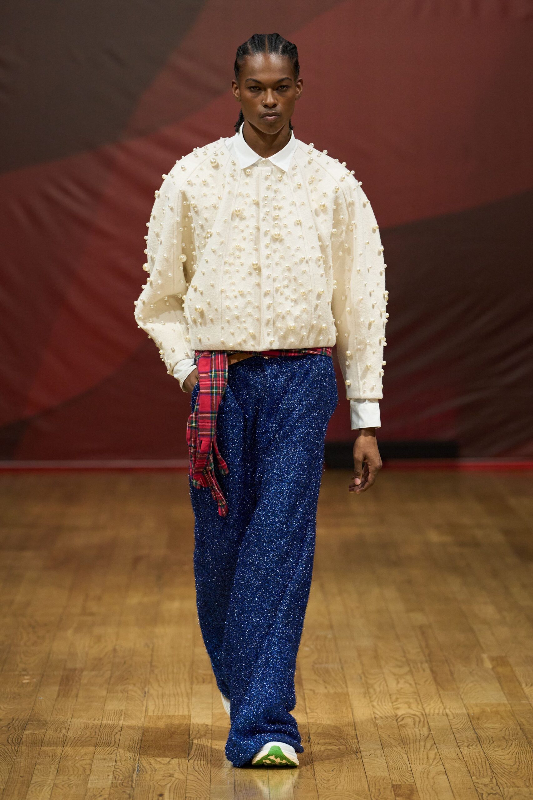 Cream button-up shirt densely studded with pearl beads, cuffs hanging long. Red tartan sash knotted low, one end dropped past the hip. Cobalt glitter-knit trousers pooling heavily over the shoes. White sneakers with a flash of green at the sole. Walking across a town hall floor after the holiday raffle is done.