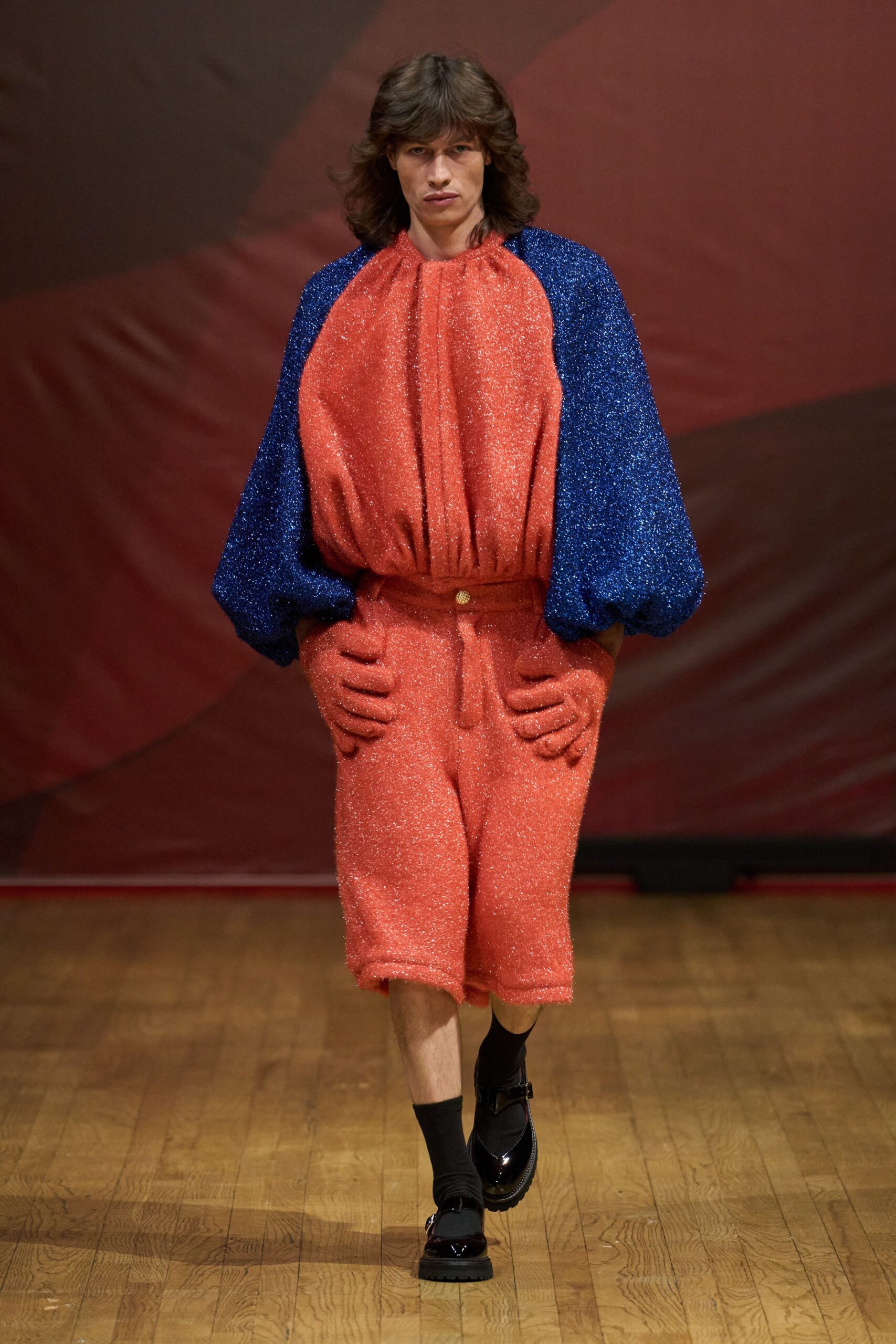 Coral glitter knit romper, gathered neckline and blue balloon sleeves. Narrow rust belt with a single round gold stud. Matching coral gloves, each hip topped with stuffed hands. Black ankle socks. Black patent Mary Janes with thick lug soles. For opening night at a puppet theater nobody takes seriously.