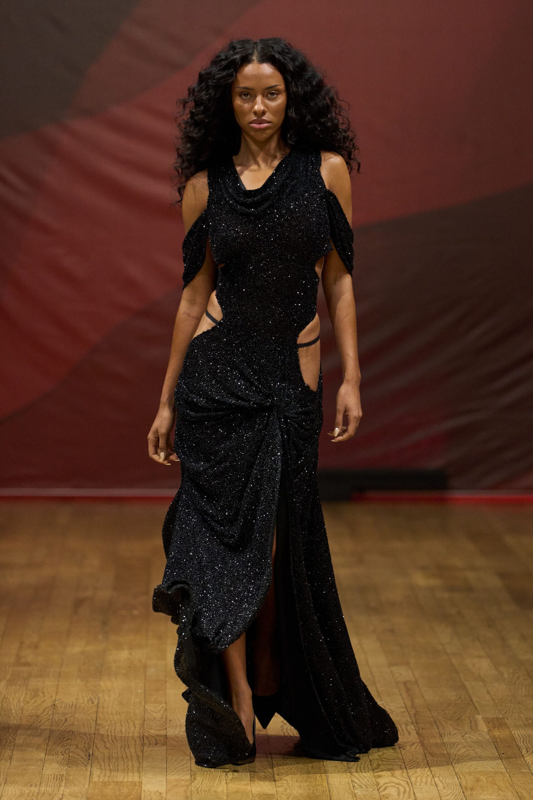 Black glitter-knit gown with a draped cowl neck and hip cutouts. Detached shoulder drapes hanging like short capelets from each arm. Black pointed pumps under the pooled front twist. Whatever you wear to cross a hotel ballroom after midnight awards.