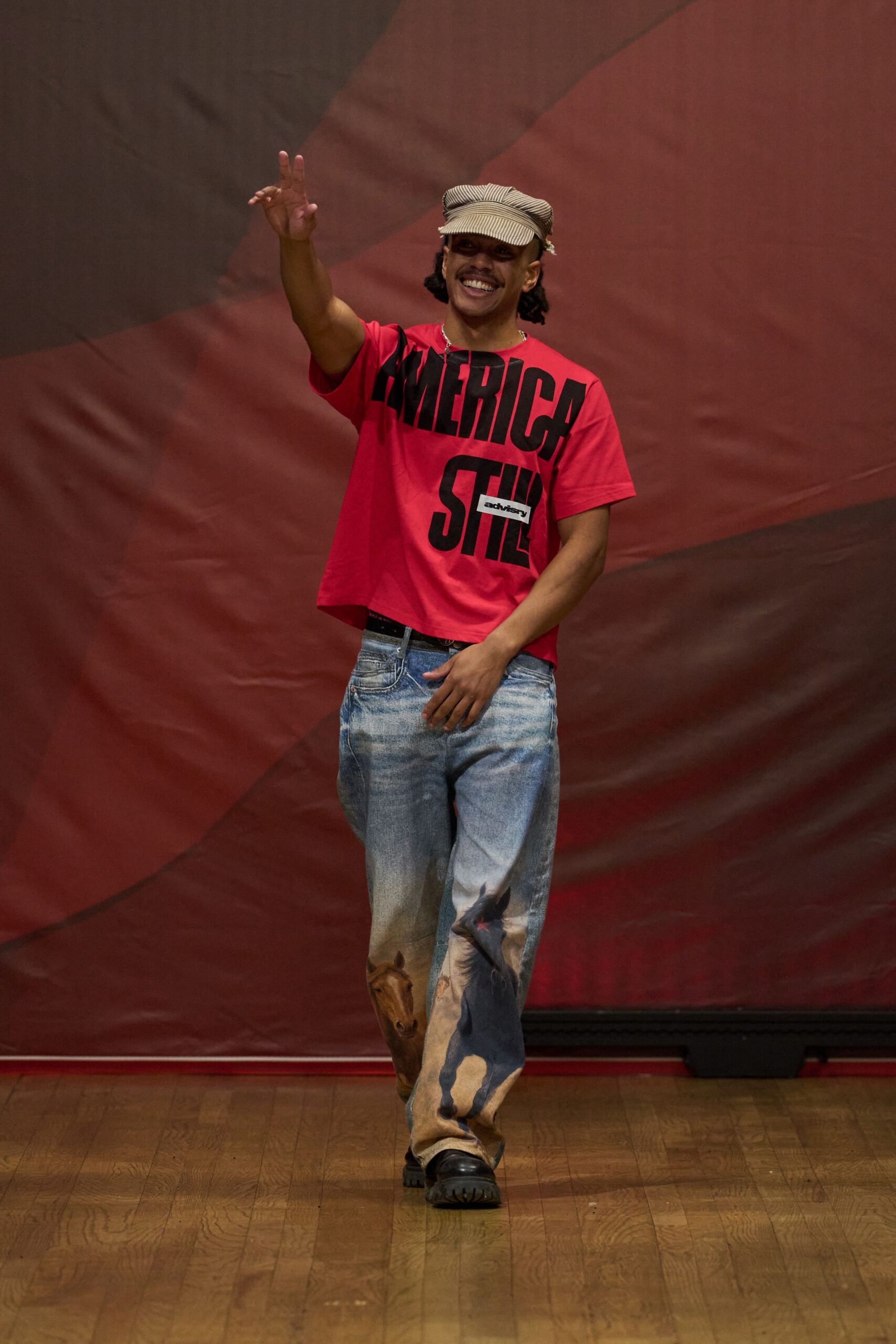 Tan railroad-stripe cap with a softly crushed crown. Boxy scarlet cotton tee, oversized black block letters swallowing the chest. Light-wash jeans airbrushed with running horses up both shins. Black lug-sole shoes peeking bluntly from the hem. County fair grandstand, waving at someone by the livestock pens.
