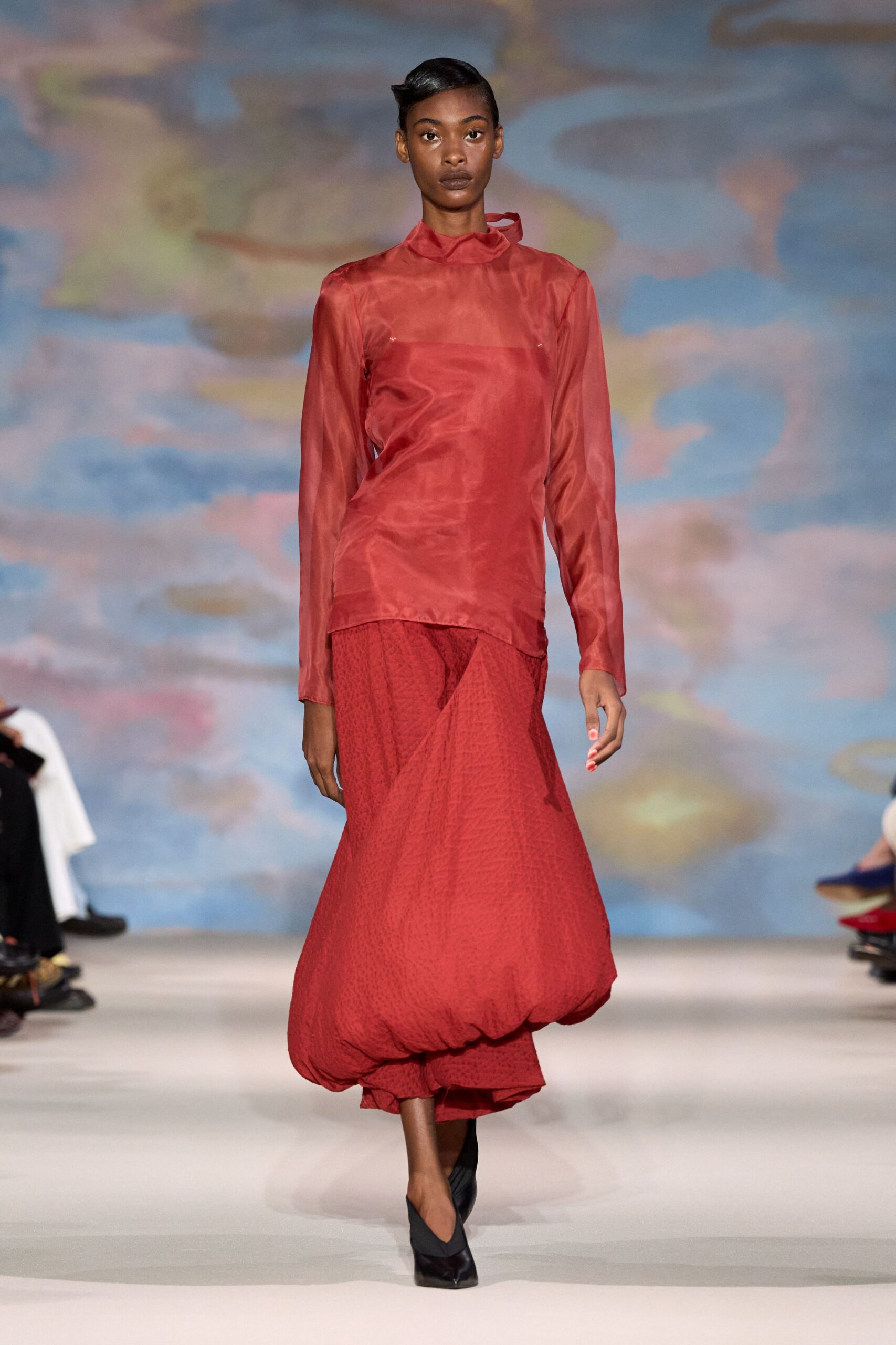 Sheer coral organza top with a twisted funnel neck and visible bandeau. Carmine textured skirt swelling into a dropped bubble hem. Black pumps with sharply pointed toes. Walking out to the hotel terrace just before the storm finally breaks.