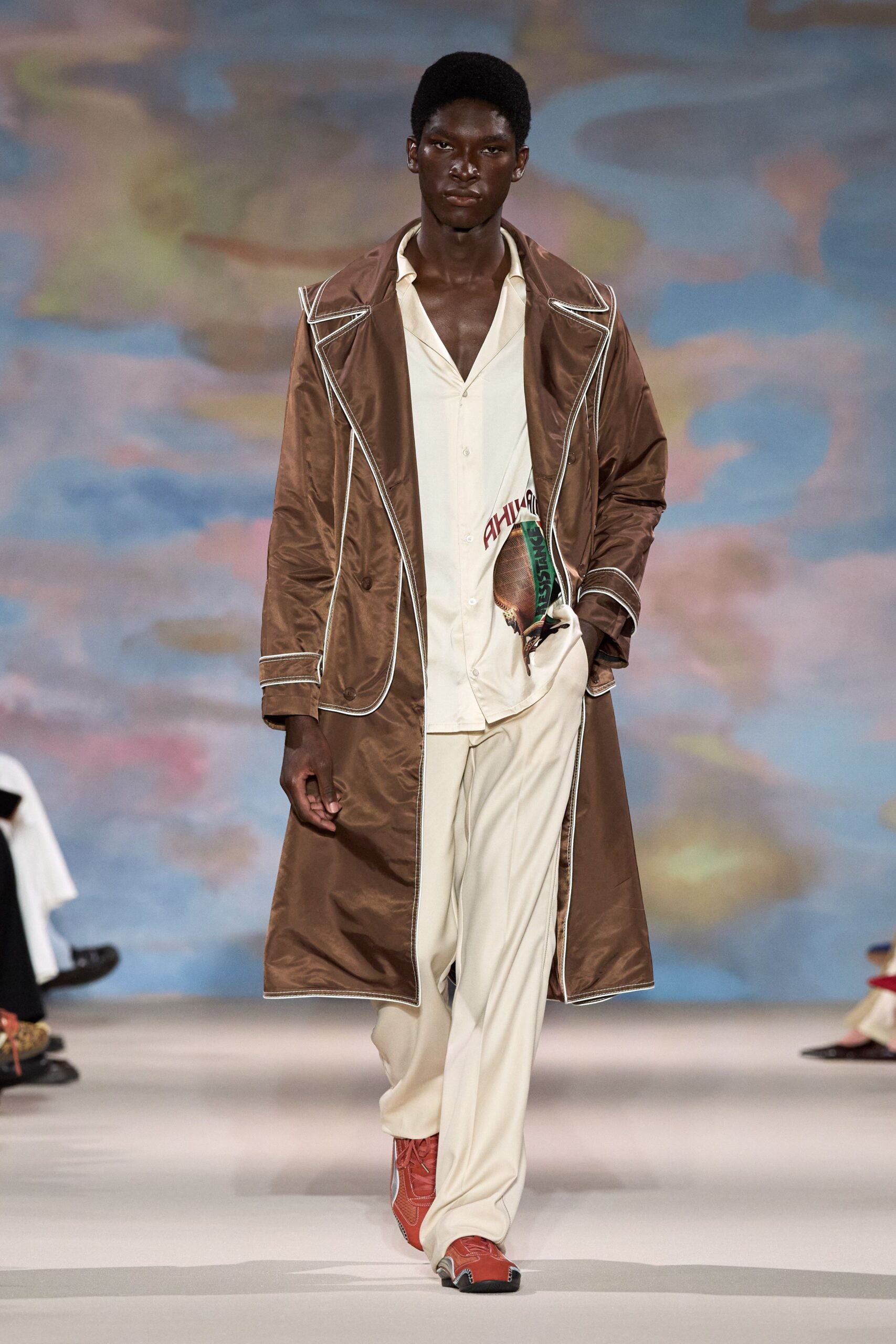 Chestnut nylon trench, oversized lapels traced in white piping. Cream camp-collar shirt hanging open, one low button still fastened. Cream fluid trousers breaking softly over the shoe. Red leather runners with silvered toe caps. Crossing the ferry deck after sunset, wind pushing the coat open.