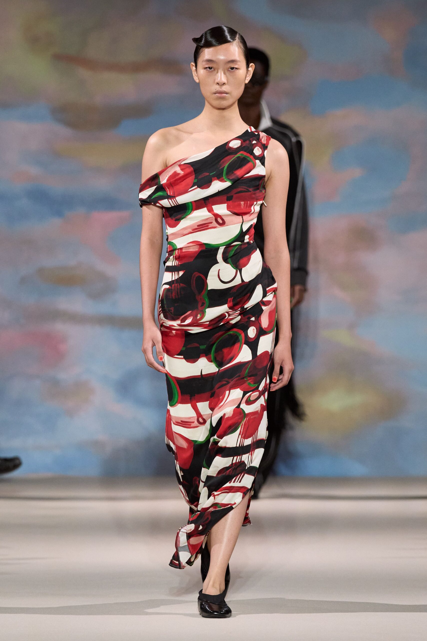 One-shoulder jersey column dress, ruched through the hip in red-black abstract print. Black mesh flats with glossy rounded cap toes. Walking into a hotel bar still carrying the glare of late afternoon.