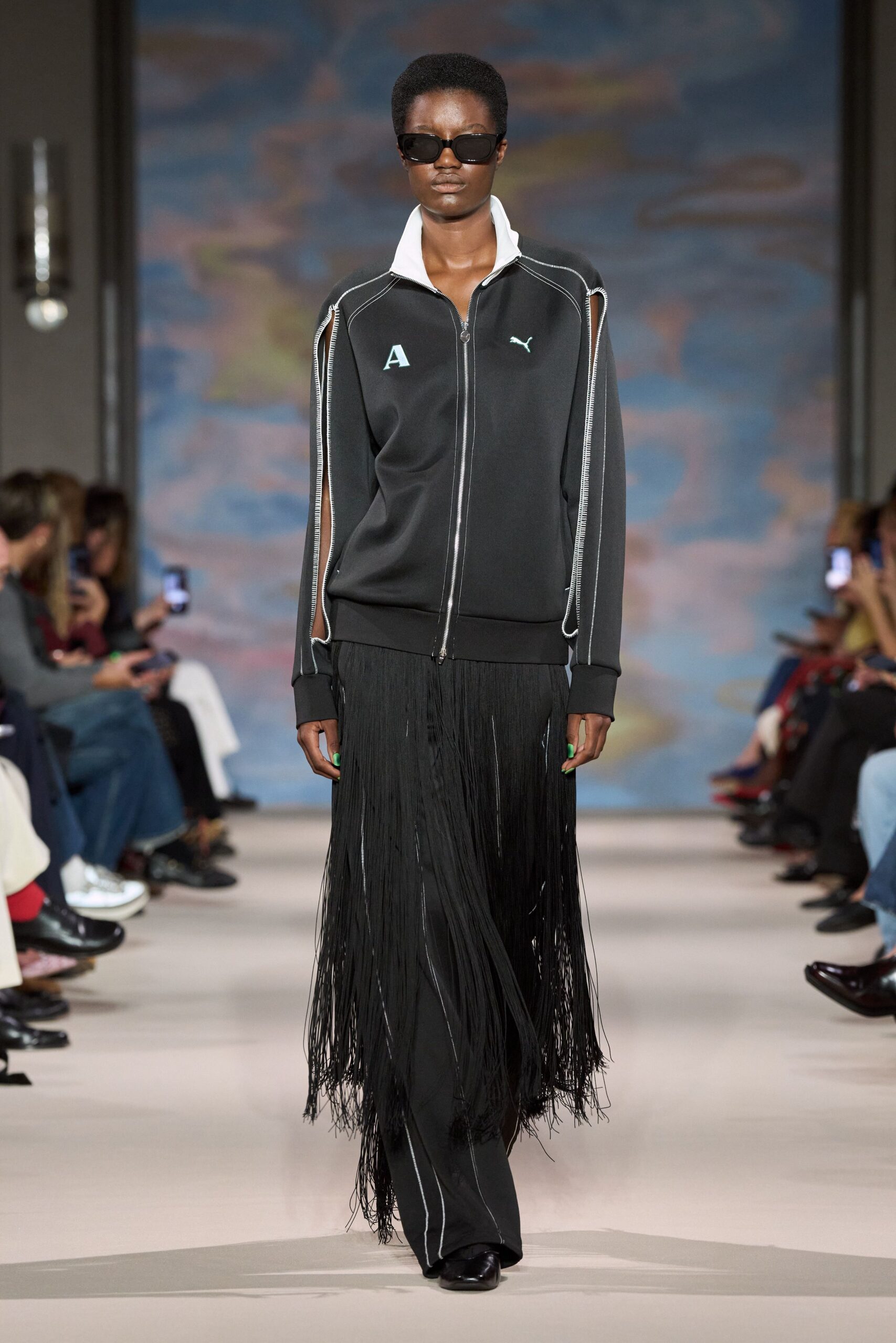 Black wraparound sunglasses. Black zip-front track jacket, white stand collar and unzipped sleeve seams. Dense black fringe overskirt swaying from the hips to mid-calf. Black wide-leg trousers with thin white seam piping. Square-toe black shoes. Walking across the station concourse after missing the last easy train.
