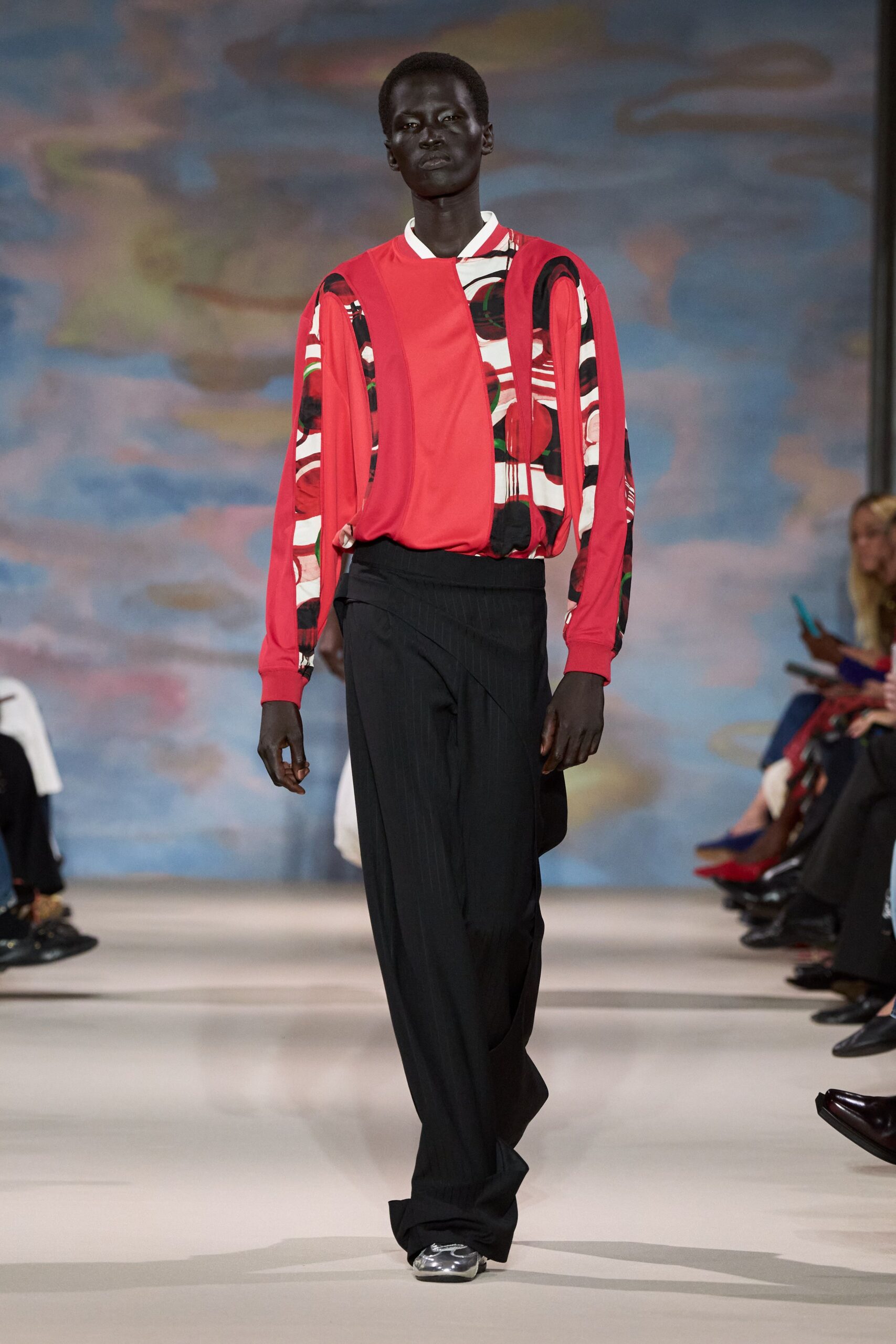 Tomato-red blouse spliced with white graphic panels and gathered sleeves. Black pinstriped wrap-front trousers, waistband crossing low and slightly twisted. Silver trainers peeking from the pooled hem. Walking across an airport atrium before the first coffee kiosk opens.