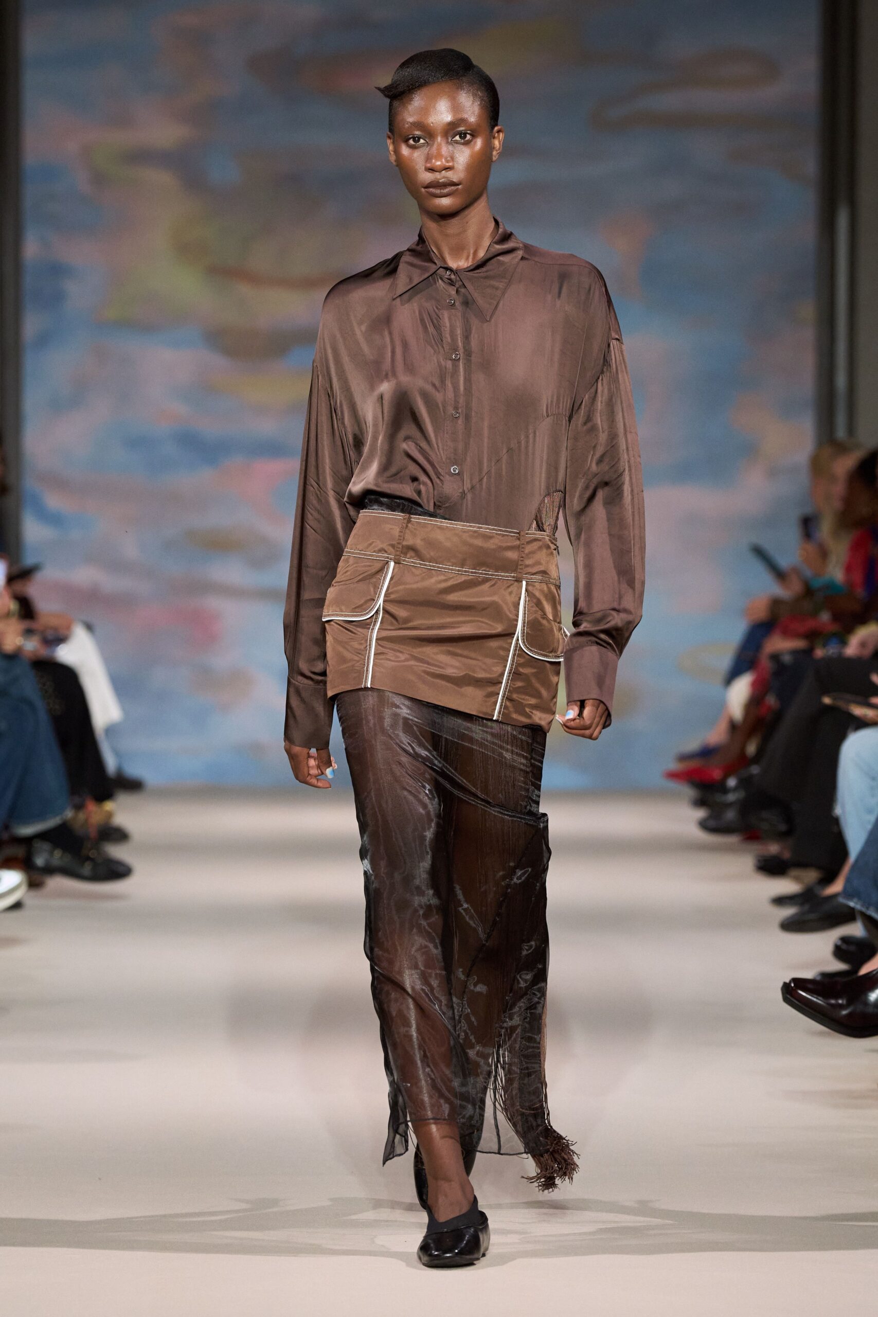 Chocolate satin blouse with dropped shoulders and a pointed collar. Taupe nylon overskirt, white piping tracing the patch pockets. Sheer espresso skirt streaked with cloudy marbling, fringe dragging at one corner. Black flats with squared toes. Walking back from the launderette while the pavement is still drying.