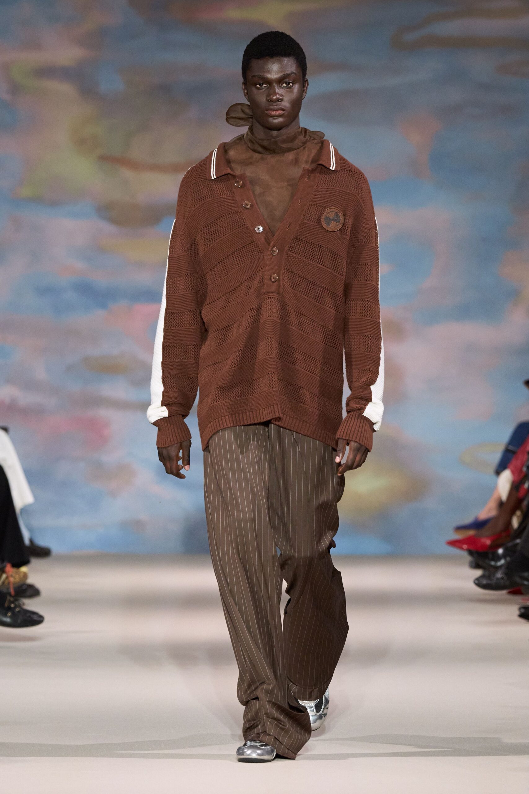 Sheer brown scarf knotted close, one end floating behind the neck. Chestnut open-knit polo, deep henley placket and striped collar tips. Loose cocoa pinstripe trousers pooling over the instep. Silver trainers with rounded white toes. Walking the seafront at dusk after the heat finally broke.