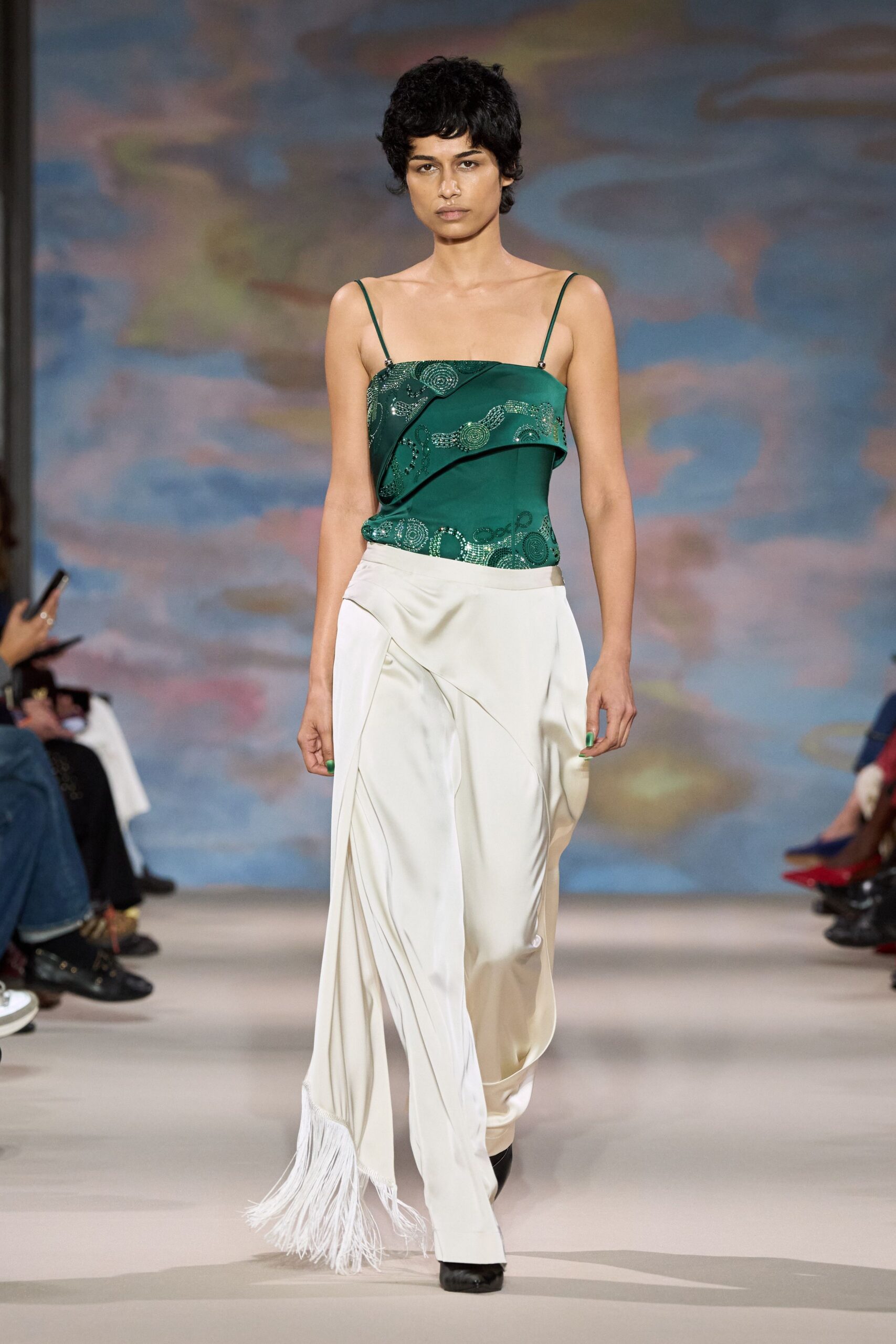 Emerald camisole top with silver beaded swirls and a folded asymmetric panel. Ivory satin trousers cut loose, one hem trailing into a fringe sweep. Black pointed boots. Crossing a hotel lobby at dawn with yesterday's event still on you.