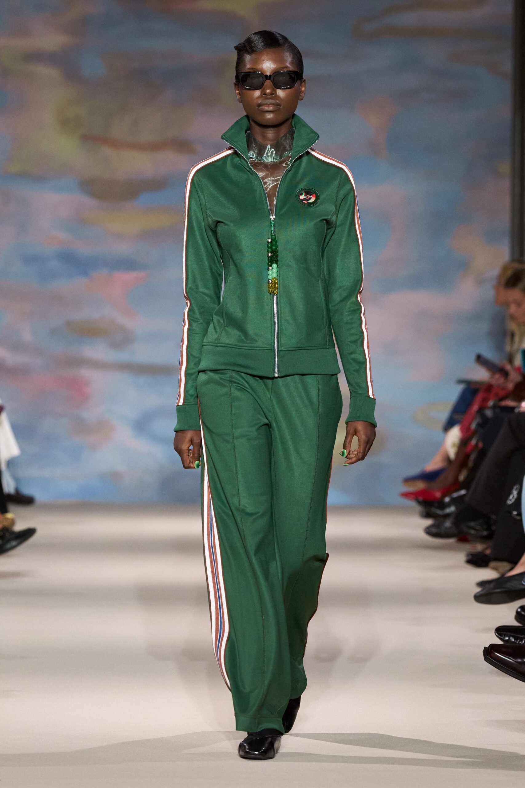 Narrow black wraparound sunglasses. Sheer smoke turtleneck with a wet-look crinkled surface. Forest green zip track jacket, striped sleeves and a beaded pull. Matching flared track pants with side stripes and front seam lines. Black square-toe shoes. Walking across a holiday camp forecourt before the pool opens.