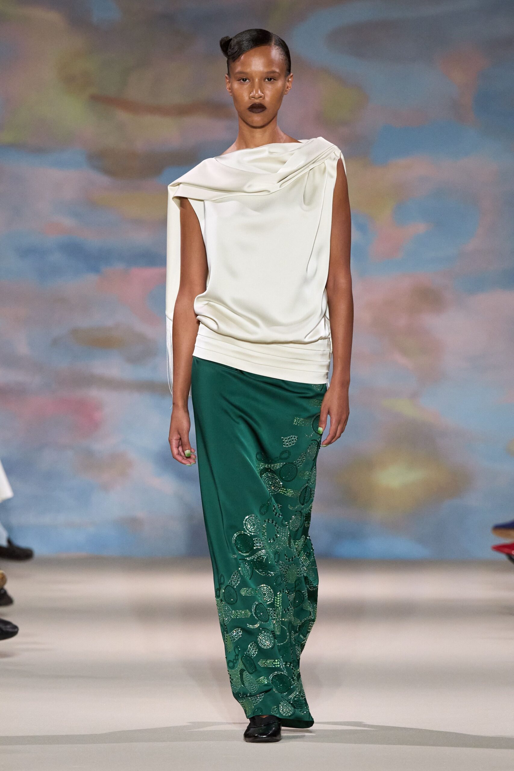 Ivory satin top with a folded bateau neckline and hanging side panel. Deep green column skirt traced with swirling beadwork near the hem. Black patent flats, square at the toe. Museum fundraiser before dinner, standing near the painted mural wall.