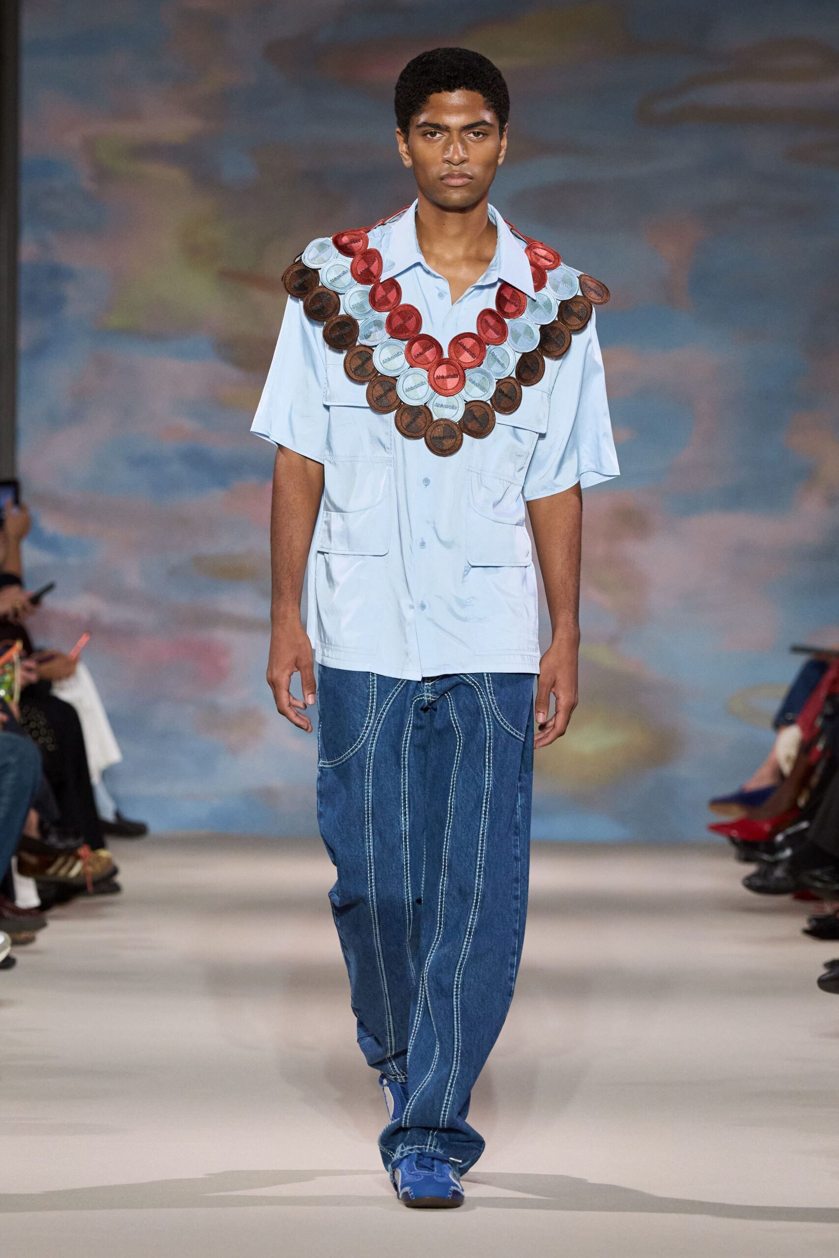 Oversized bib of stitched circular patches in red, pale blue and brown. Powder-blue camp-collar shirt with oversized flap pockets and a soft drape. Dark indigo wide jeans traced with looping white topstitch curves. Blue sneakers peeking from the pooled hem. Walking to a beach fair with prize tokens still hanging round your neck.