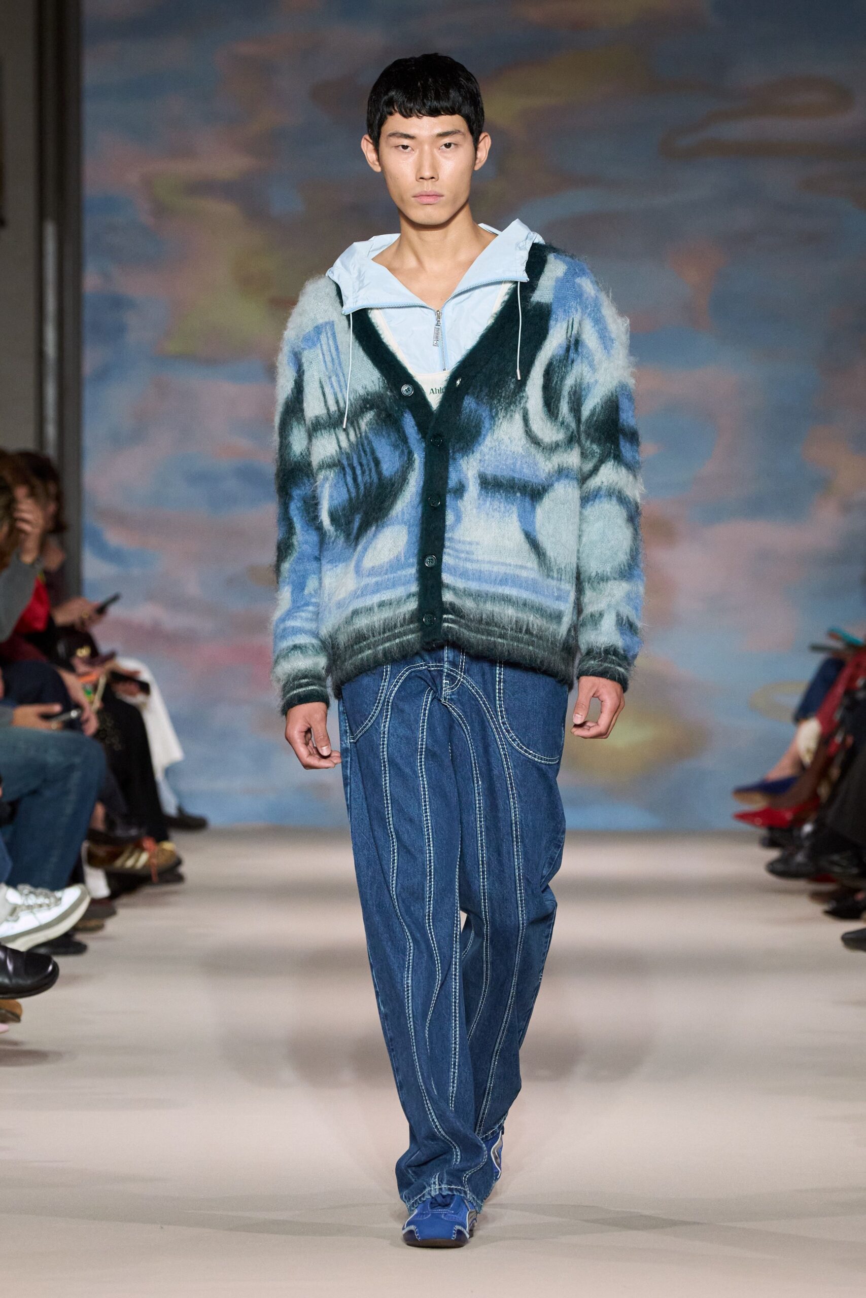 Powder-blue nylon hooded top, zip left open below the throat. White tank peeking through the V. Fuzzy brushed cardigan in clouded blue and black, dark rib placket. Wide indigo jeans mapped with looping white topstitch seams. Blue shoes with white whipstitched toes. Walking the seafront early, wind pushing mist off the water.