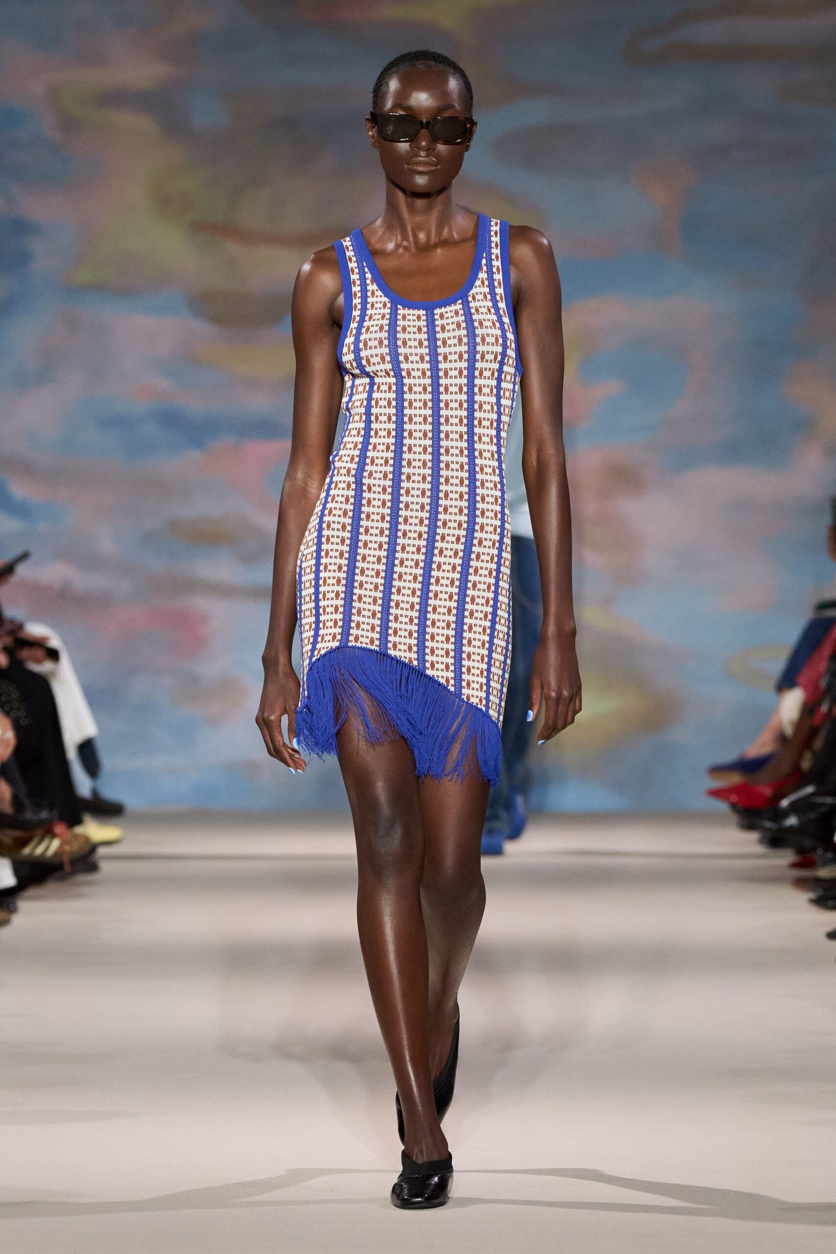 Narrow black rectangular frames. Sleeveless cream knit mini gridded in tan, traced with cobalt vertical bands. Asymmetric cobalt fringe swinging from the dipped front hem. Black square-toe pumps with a soft crushed vamp. Walking the boardwalk before noon, wind lifting the arcade dust.