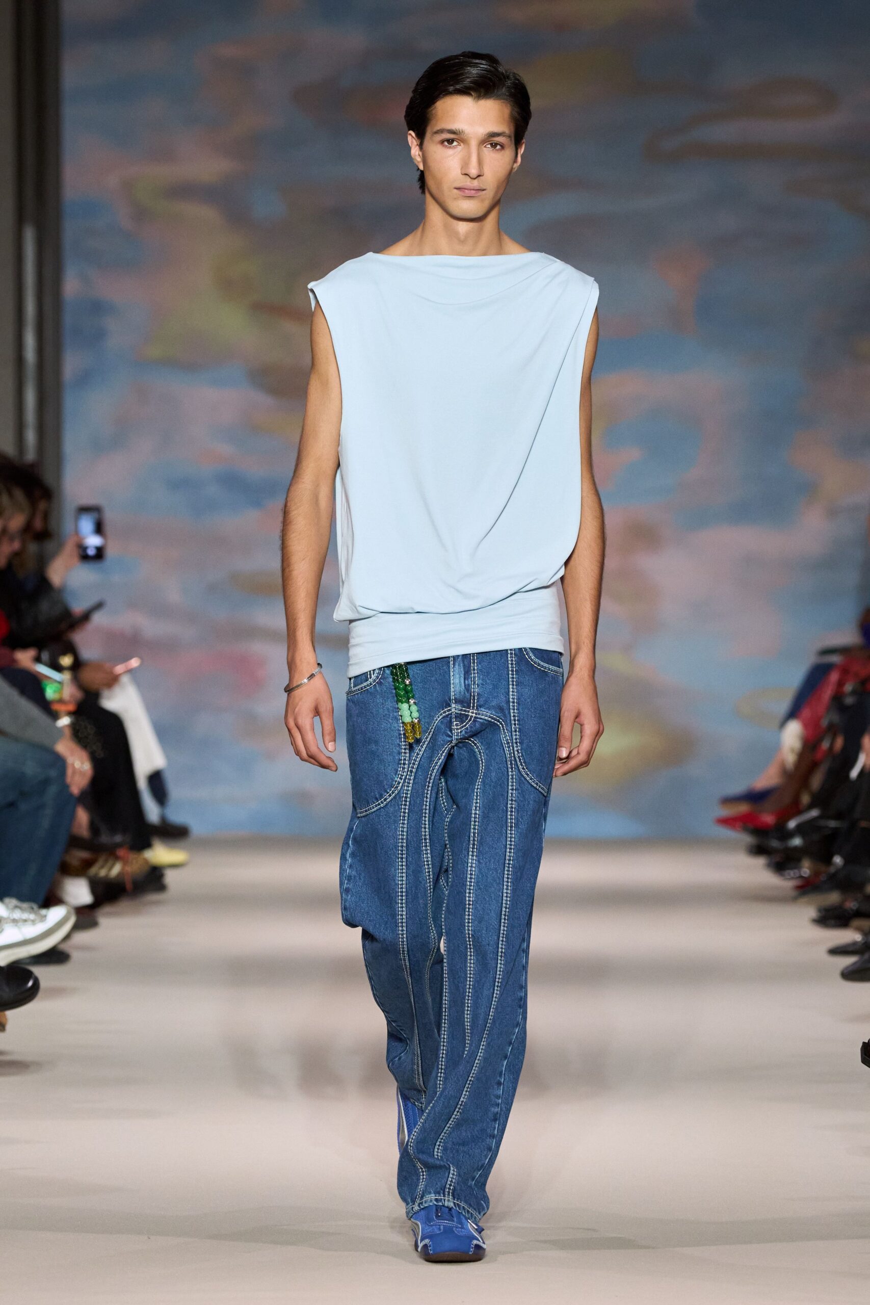 Powder-blue jersey top draped from a boat neck into a wrapped waist. Slim silver bangle. Indigo denim trousers traced with looping contrast topstitch and a beaded side tassel. Blue square-toe shoes peeking under the hem. Walking the seafront before noon, wind already lifting the cafe receipts.