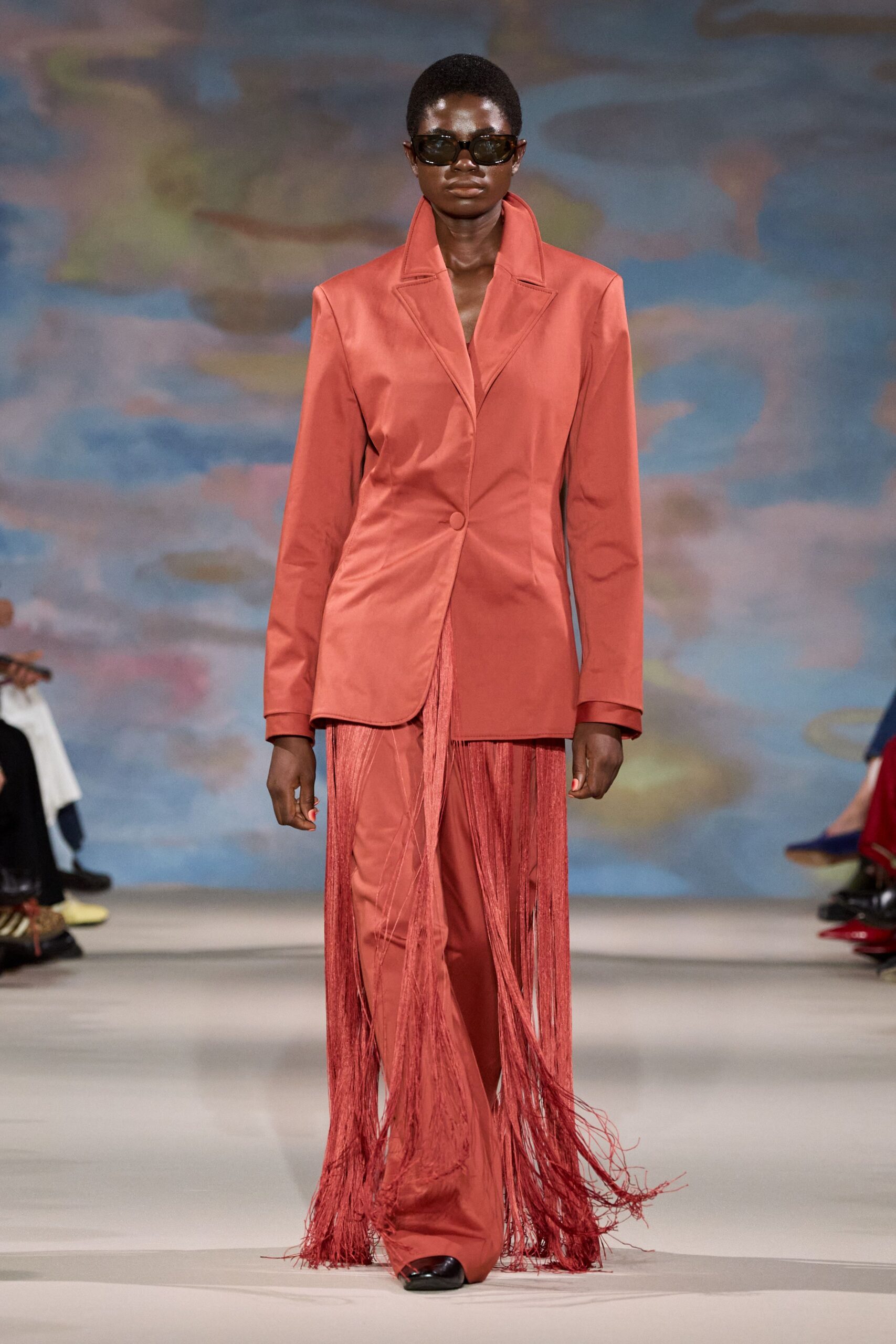 Black wraparound sunglasses with narrow smoked lenses. Coral satin blazer, high notched collar and one covered button. Matching satin trousers veiled by long swaying fringe panels. Black square-toe shoes peeking under the hem. Walking into a hotel ballroom after the florist has finished the stage.