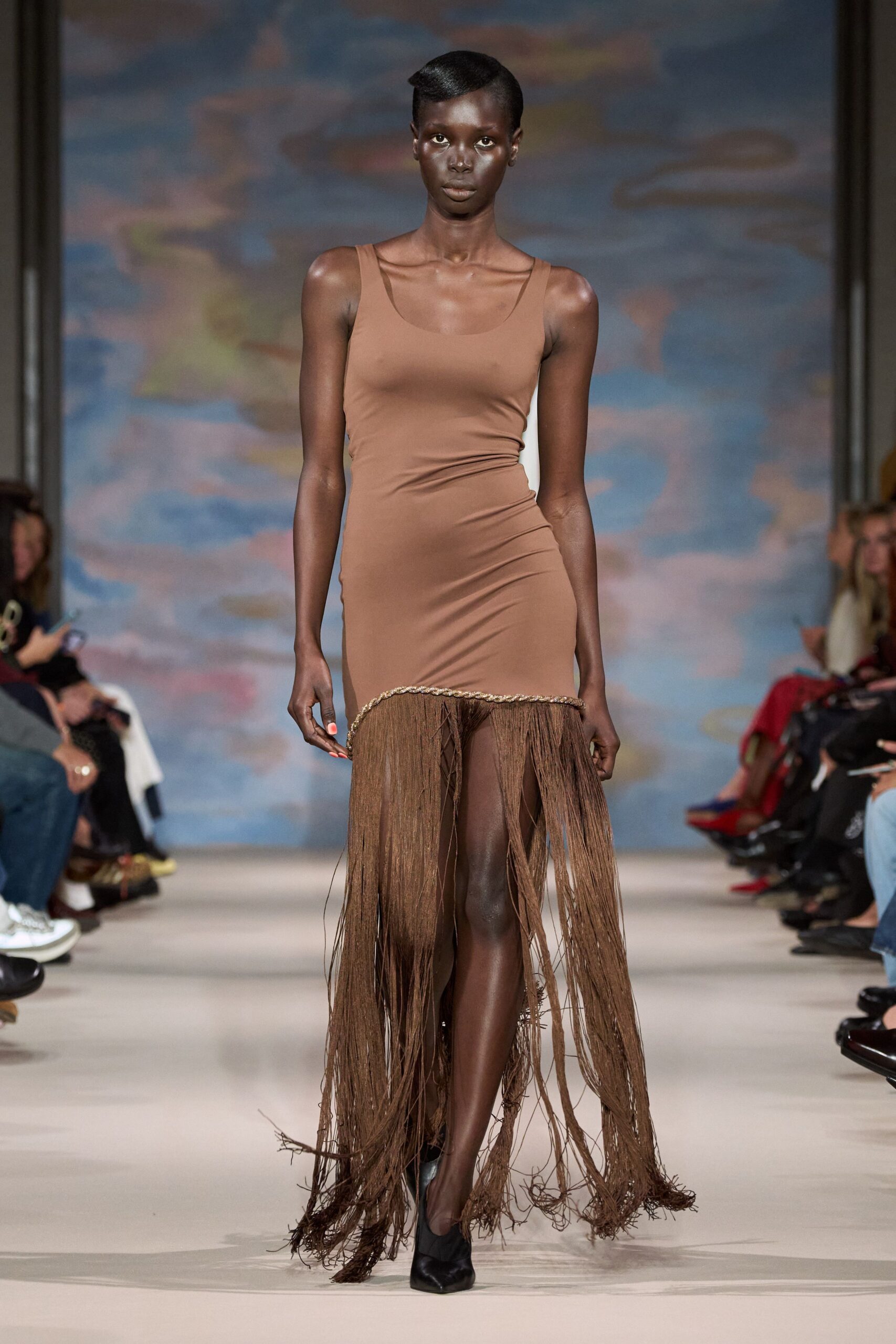Taupe stretch tank dress, side seams pulling into low hip ruching. Rope-trimmed dropped hem exploding into floor-sweeping brown tassels. Black pointed pumps. Walking across a hotel terrace after rain, hem drinking from the puddles.