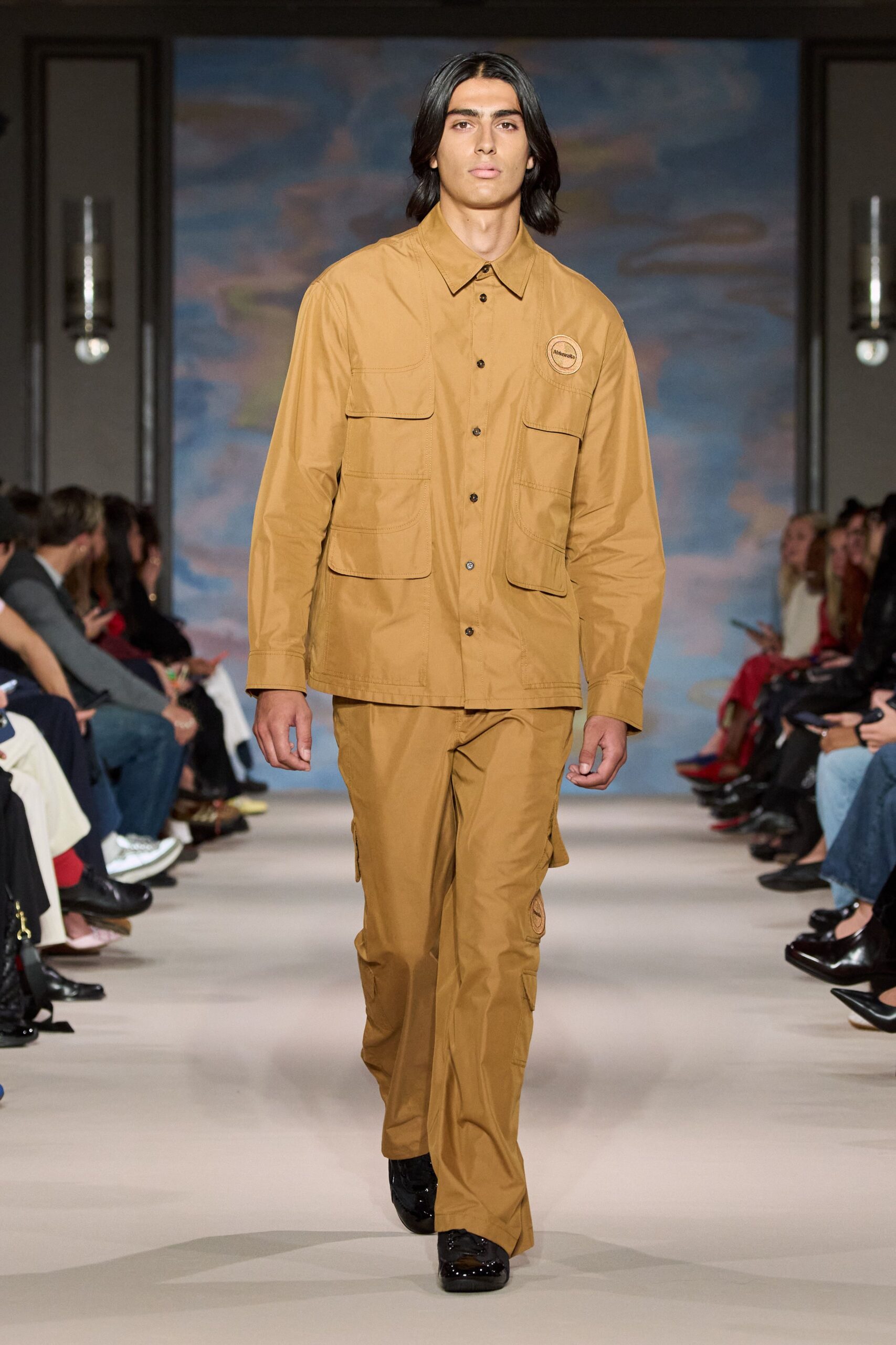 Camel cotton field jacket with four rounded flap pockets and black buttons. Matching straight cargo trousers, side pockets swelling at the thigh. Black patent shoes, square and mirror-bright. For checking fence lines after rain, when the track is still tacky.