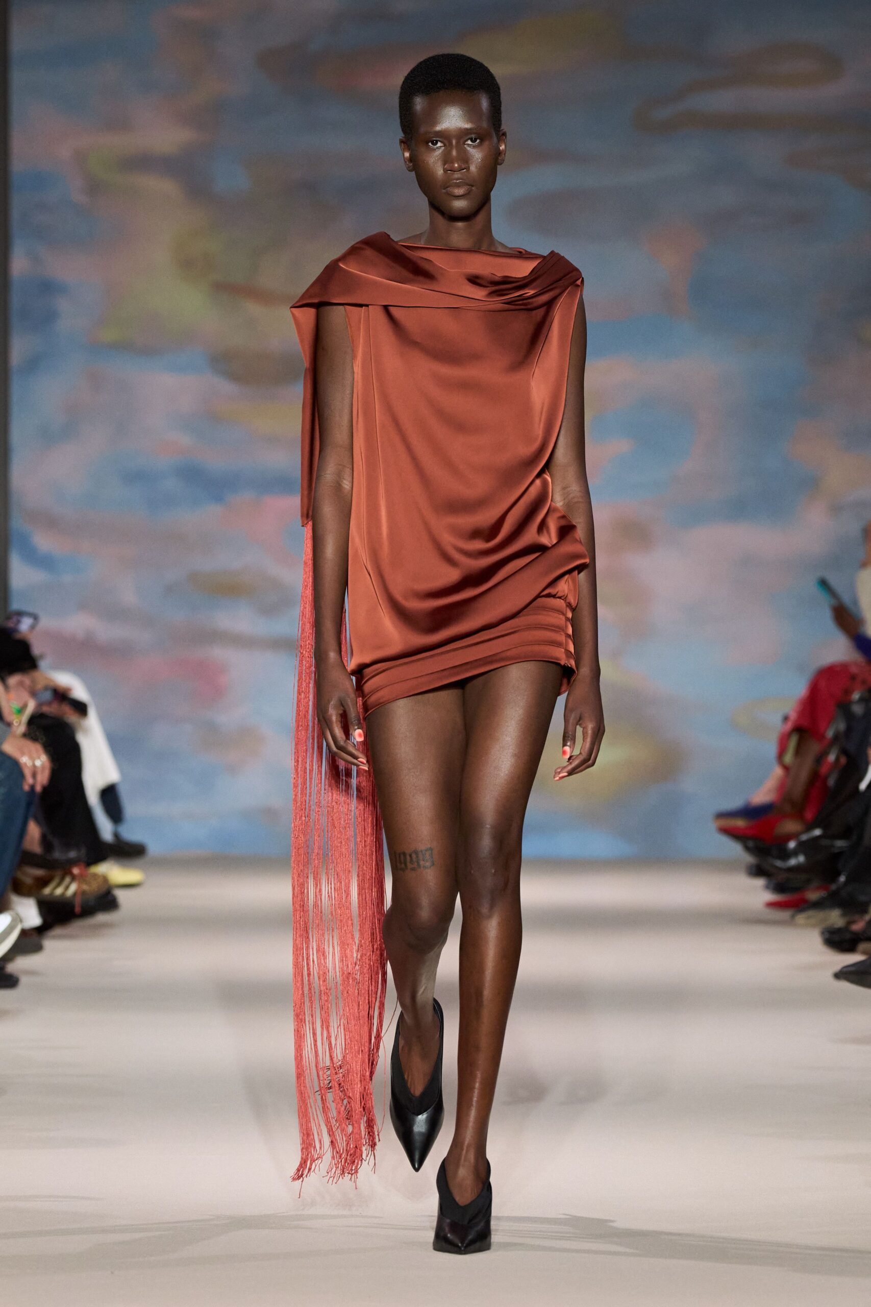 Rust satin mini dress, one shoulder spilling into a long fringe panel. Black leather pumps with sharply pointed vamps. For walking out to the terrace after sunset with your drink still full.