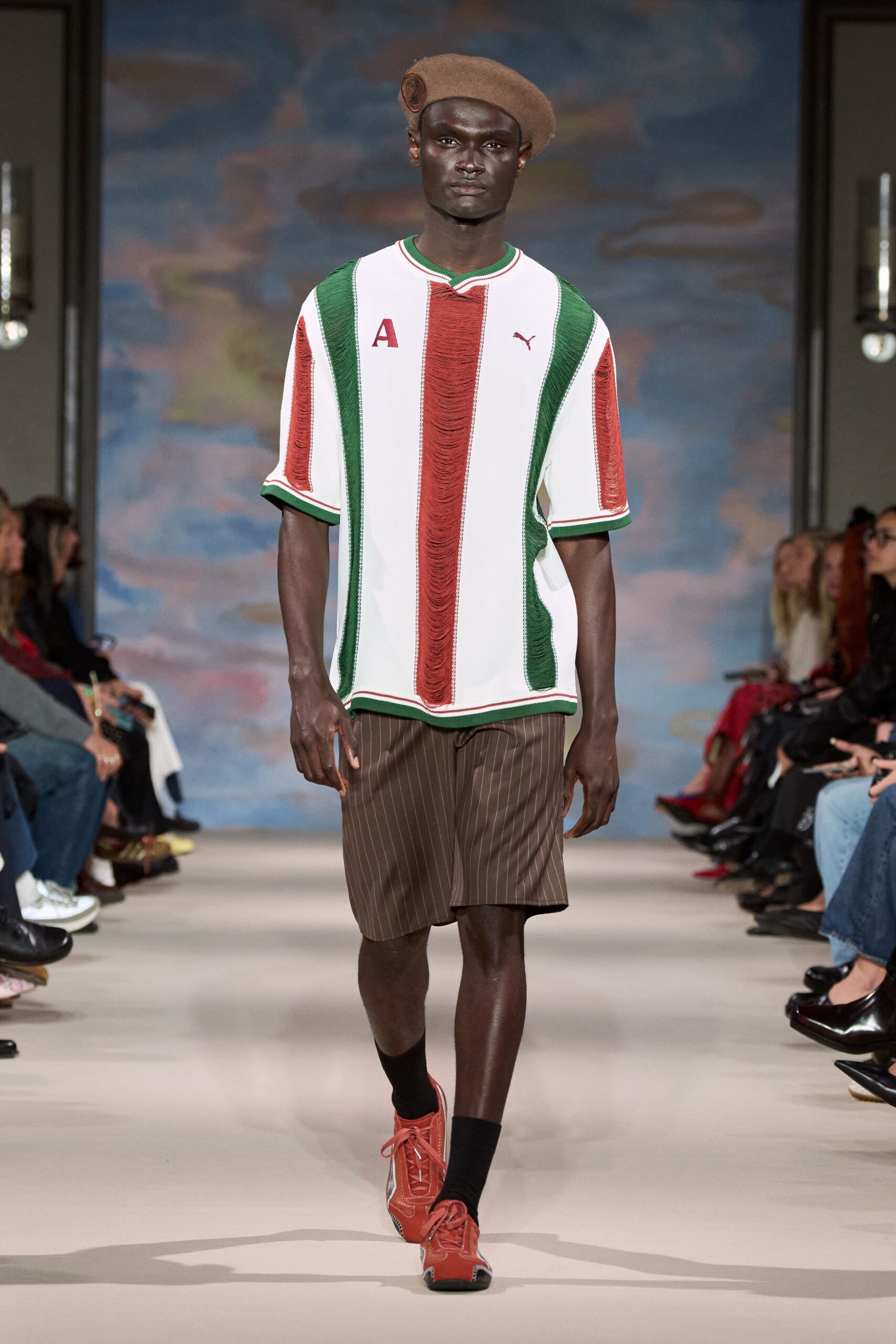 Camel felt beret with a round leather badge at one side. White football jersey cut loose, red and green laddered stripe panels. Brown pinstripe shorts, hem landing just above the knee. Black crew socks. Rust trainers with white side flashes and long laces. Walking to the corner shop after a five-a-side match, still flushed.