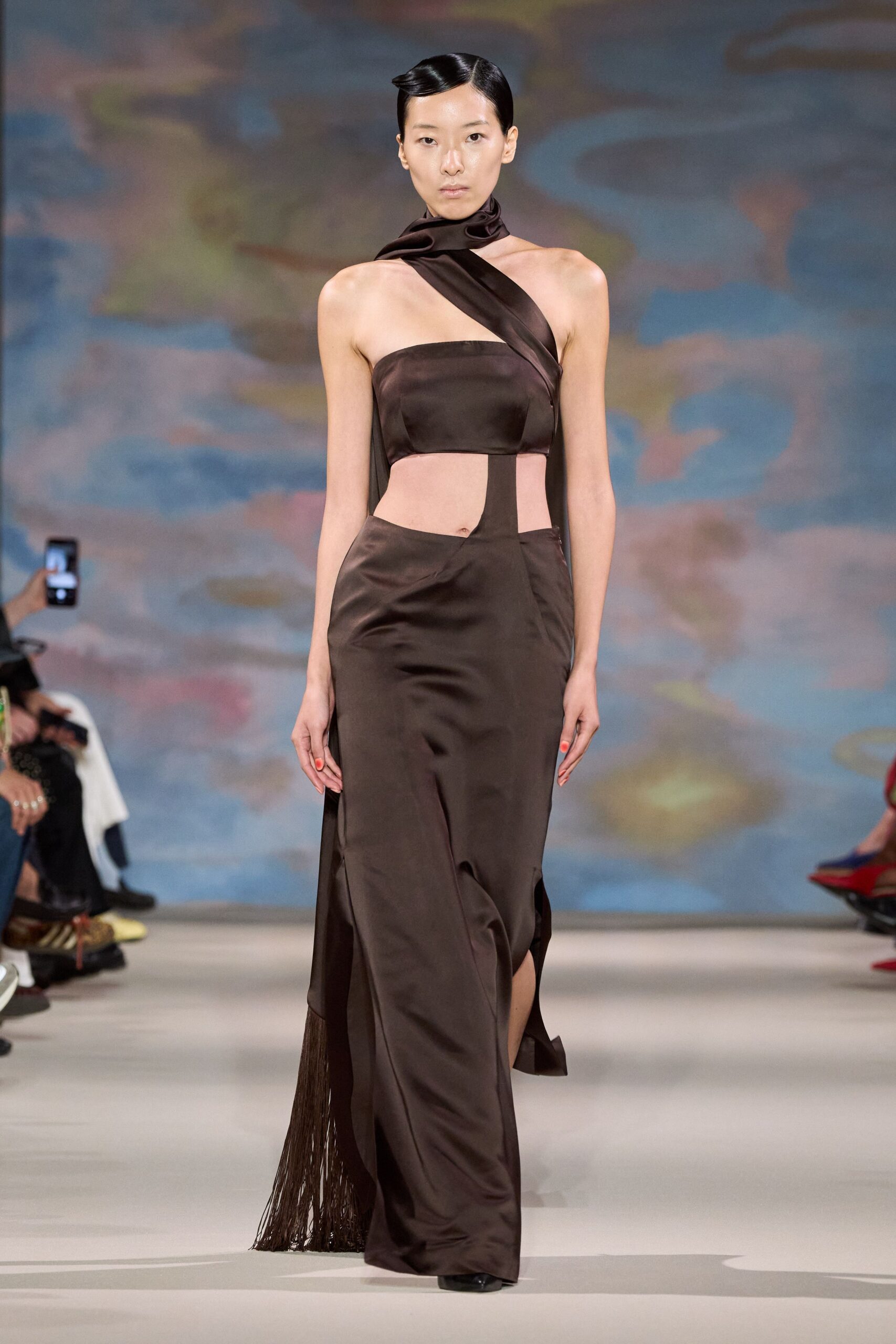 Espresso satin column gown with a bandeau bodice and carved waist cutouts. Wide satin scarf twisted at the throat, crossing into one shoulder strap. Back panel trailing into long fringe that brushes the floor behind. Black pointed toes barely breaking the hem. For crossing a hotel lobby at dawn after the wedding ended badly.