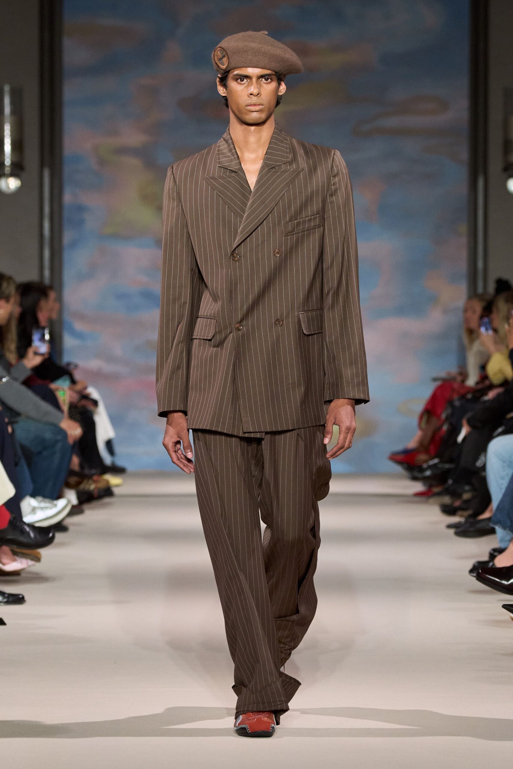 Brown wool beret with a stitched round patch at the side. Chocolate pinstripe suiting, broad shoulders and a low four-button stance. Matching pinstripe trousers breaking softly over the shoe. Rust leather shoes with white whipstitched seams at the square toe. Walking to the station with a paper ticket damp in your pocket.