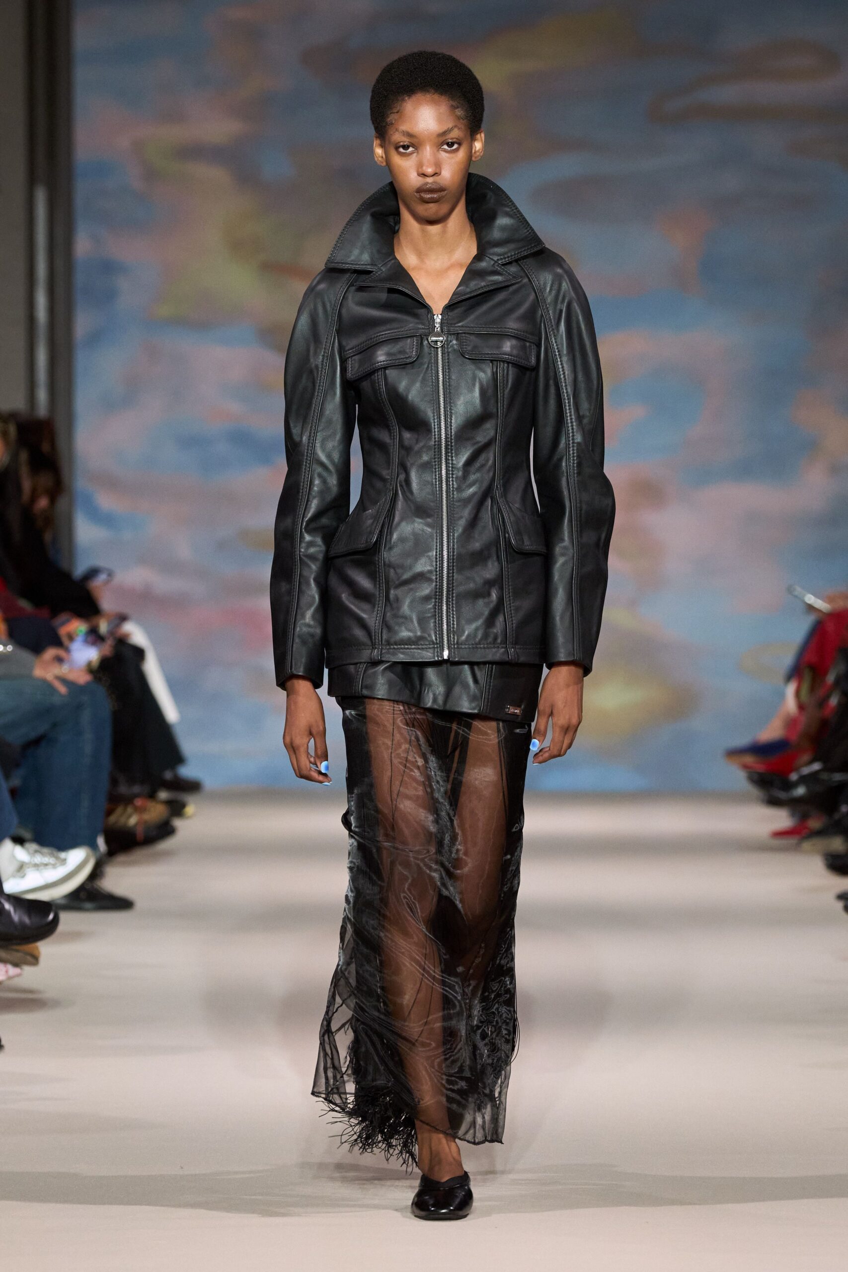 Black leather jacket sharply seamed through the waist, collar flared high. Smoke-sheer black skirt with cloudy marbling and a feathered hem. Black square-toe flats. Whatever you wear home after missing the last ferry in cold wind.
