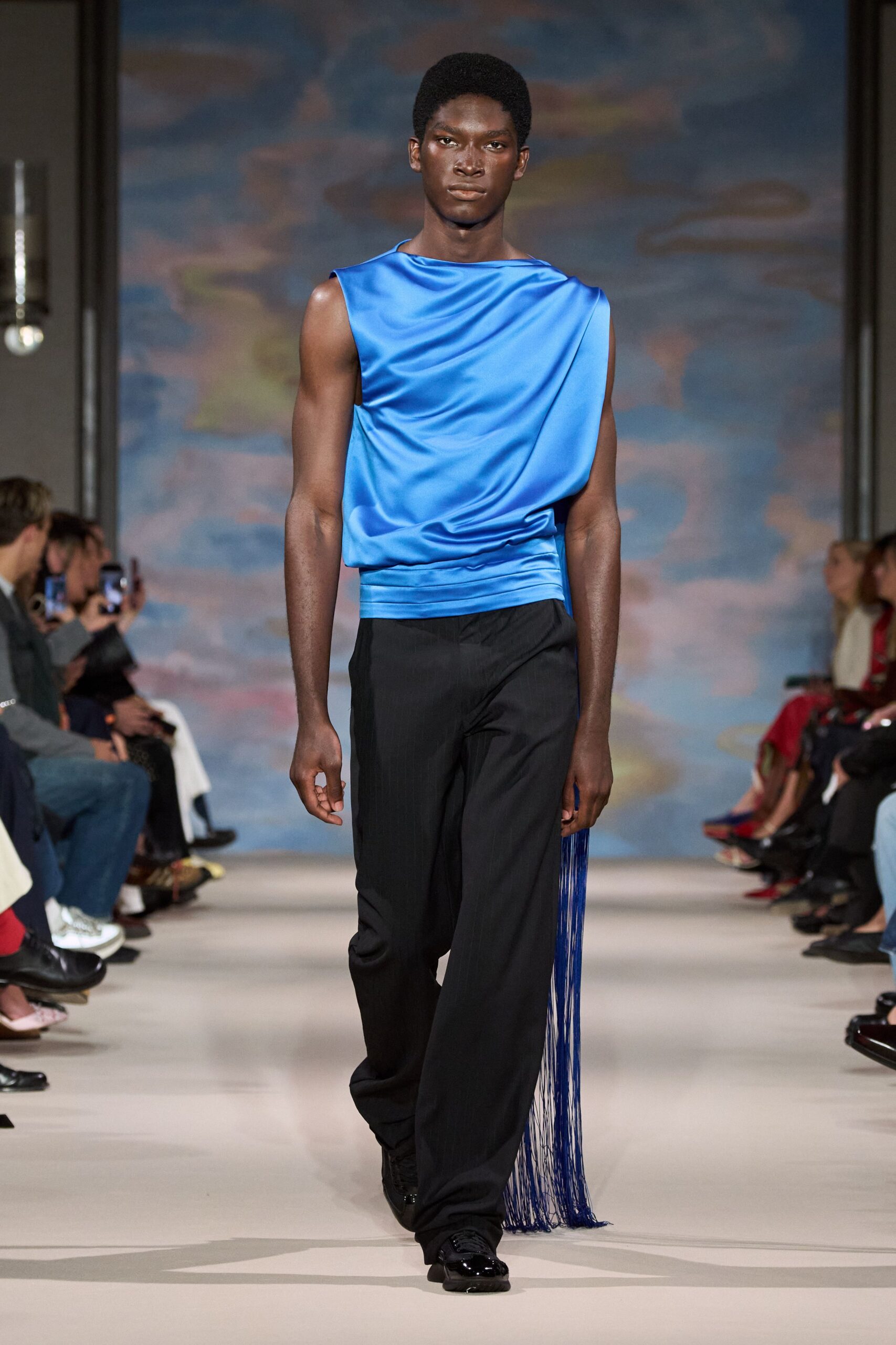 Electric-blue satin top draped diagonally, cut raw at the sleeveless edge. Black tailored trousers falling long over the shoe. Cobalt tassel panel streaming from the right hip. Black patent-toe trainers. Crossing a hotel ballroom after midnight with the sash still swinging.