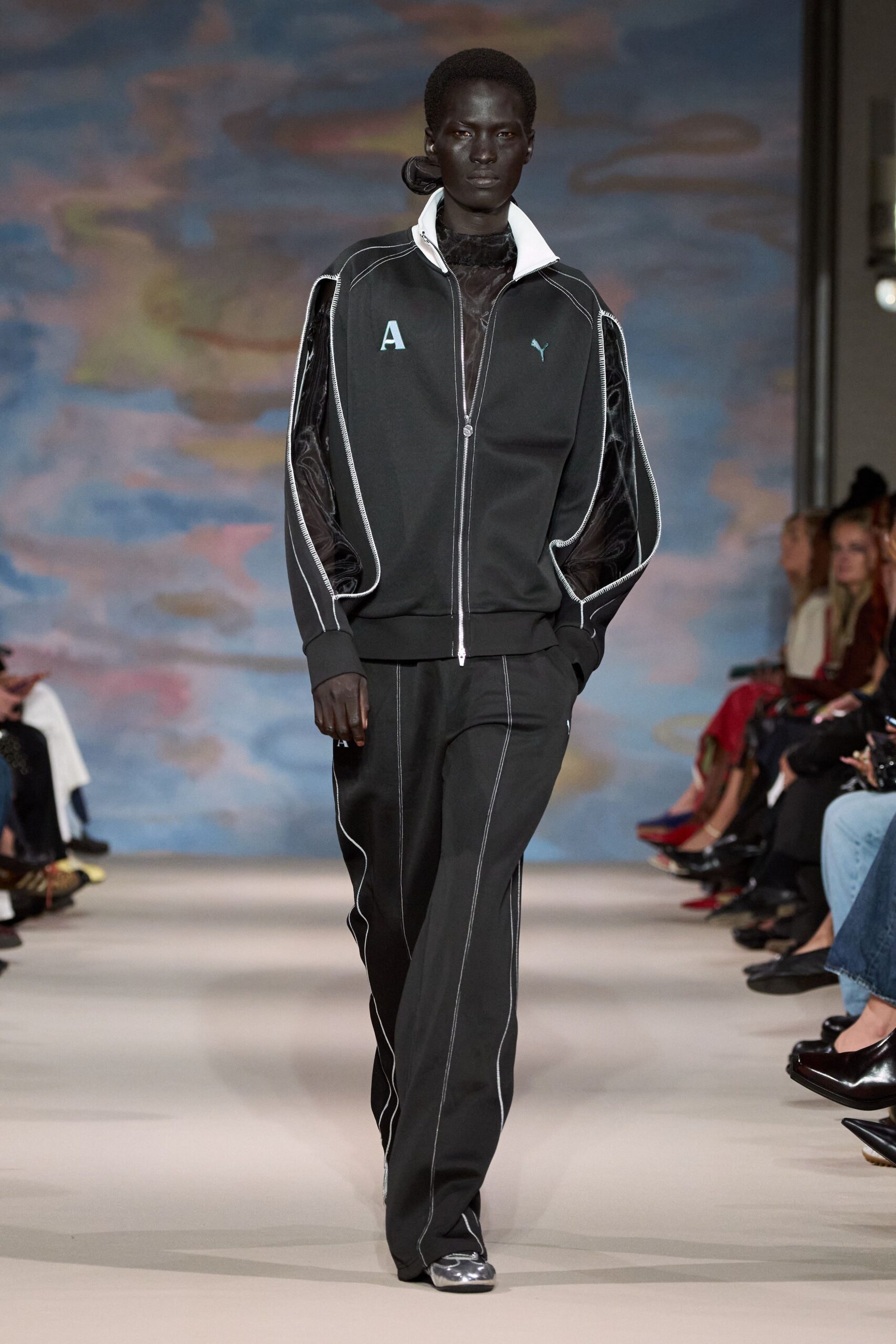 Black jersey track jacket with a white stand collar and unzipped sleeves. Sheer black top, marbled velvet curling over the throat. Matching black track pants traced with bright white topstitching. Silver shoes, mirror-bright at the square toe. Walking to the corner shop just after rain, still carrying nightclub air.