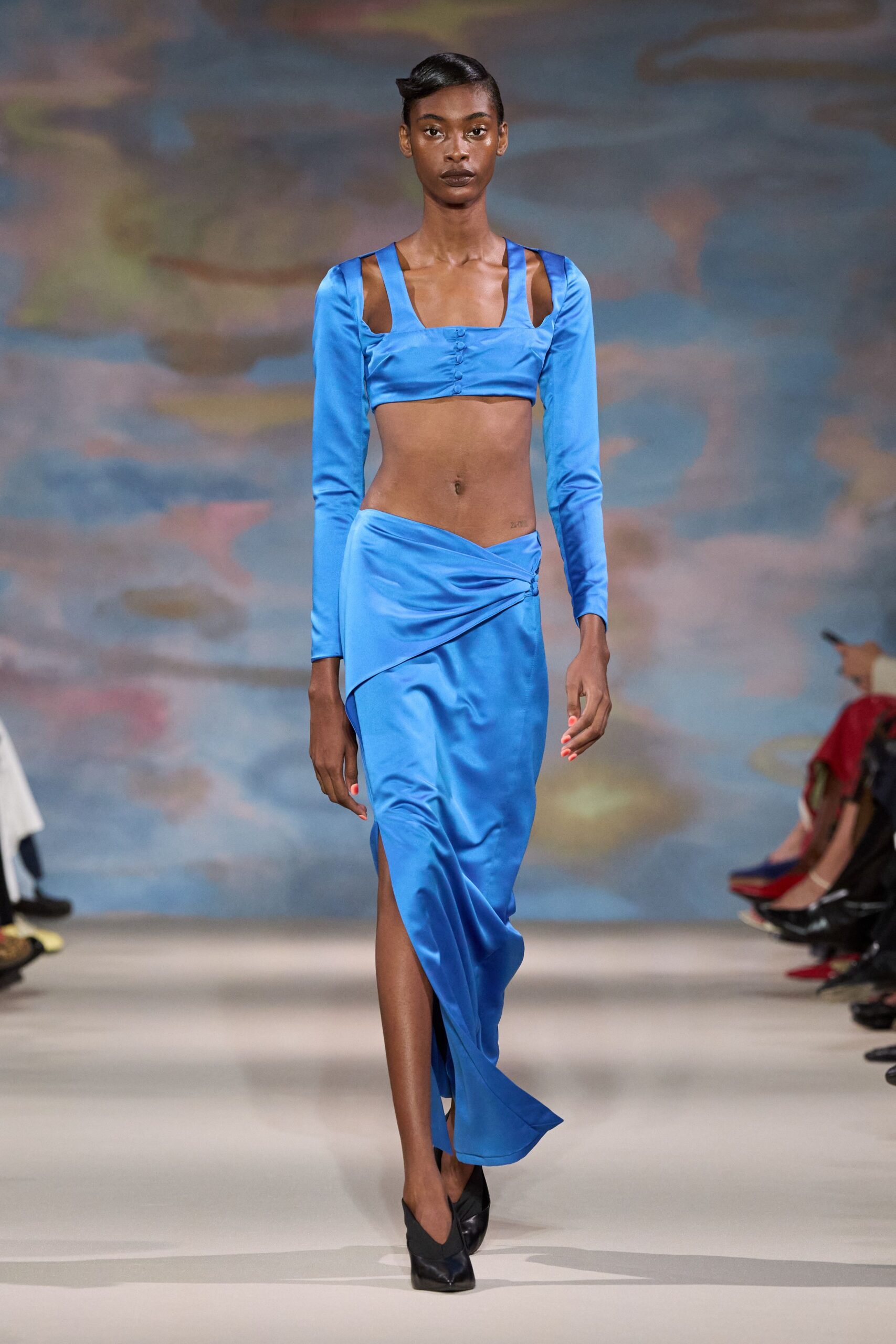Cobalt satin crop top with shoulder cutouts and tiny covered buttons. Matching bias-cut satin skirt, ruched low at one hip into a slit. Black leather pumps, sharply pointed. Walking out to the hotel terrace just before the sky turns fully dark.
