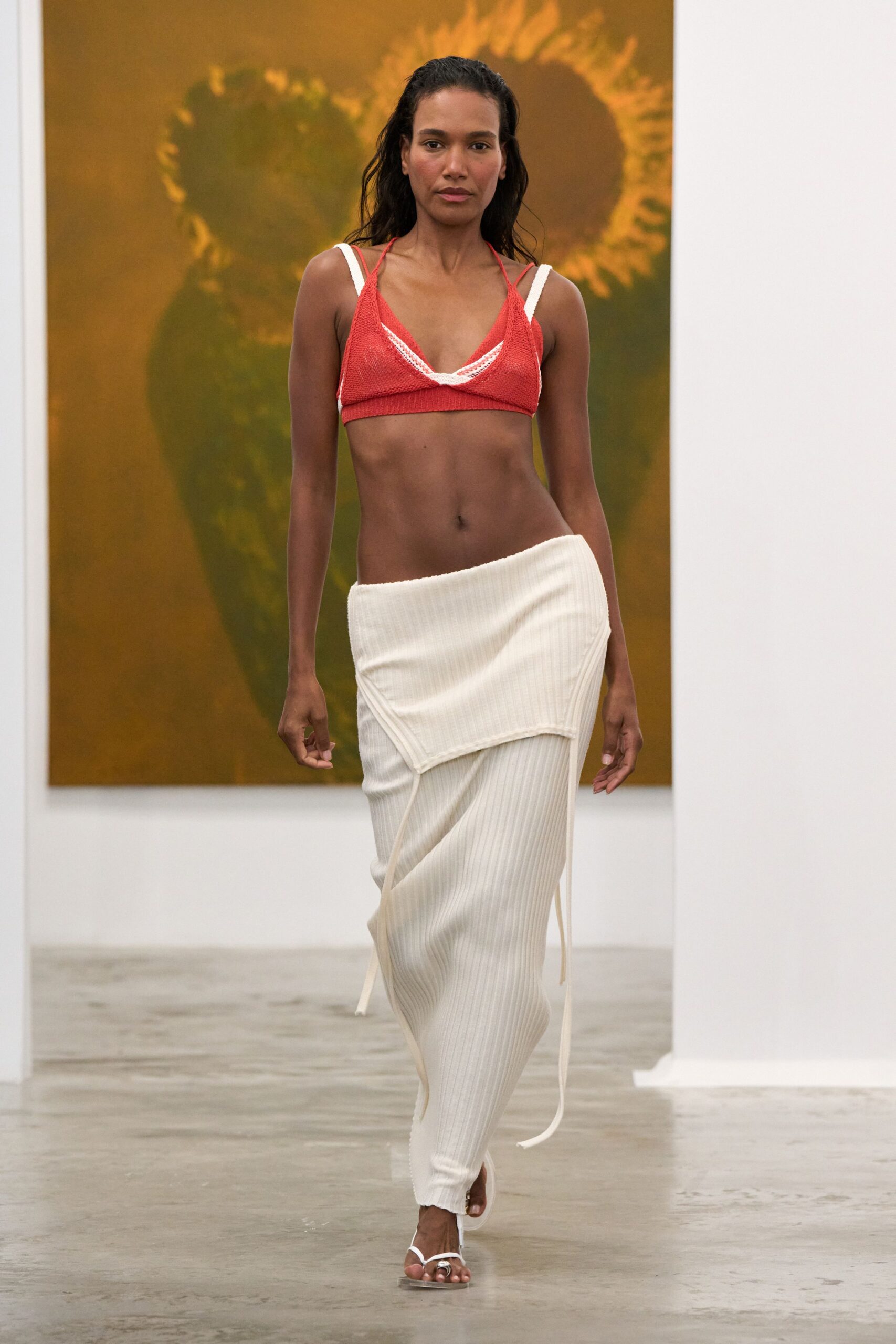 Coral crochet bralette with white binding crossing at the center. Ivory rib-knit column skirt with a slung foldover panel and dangling ties. White thong sandals with a silver toe ring. Walking through a museum atrium after swimming, hair still damp.