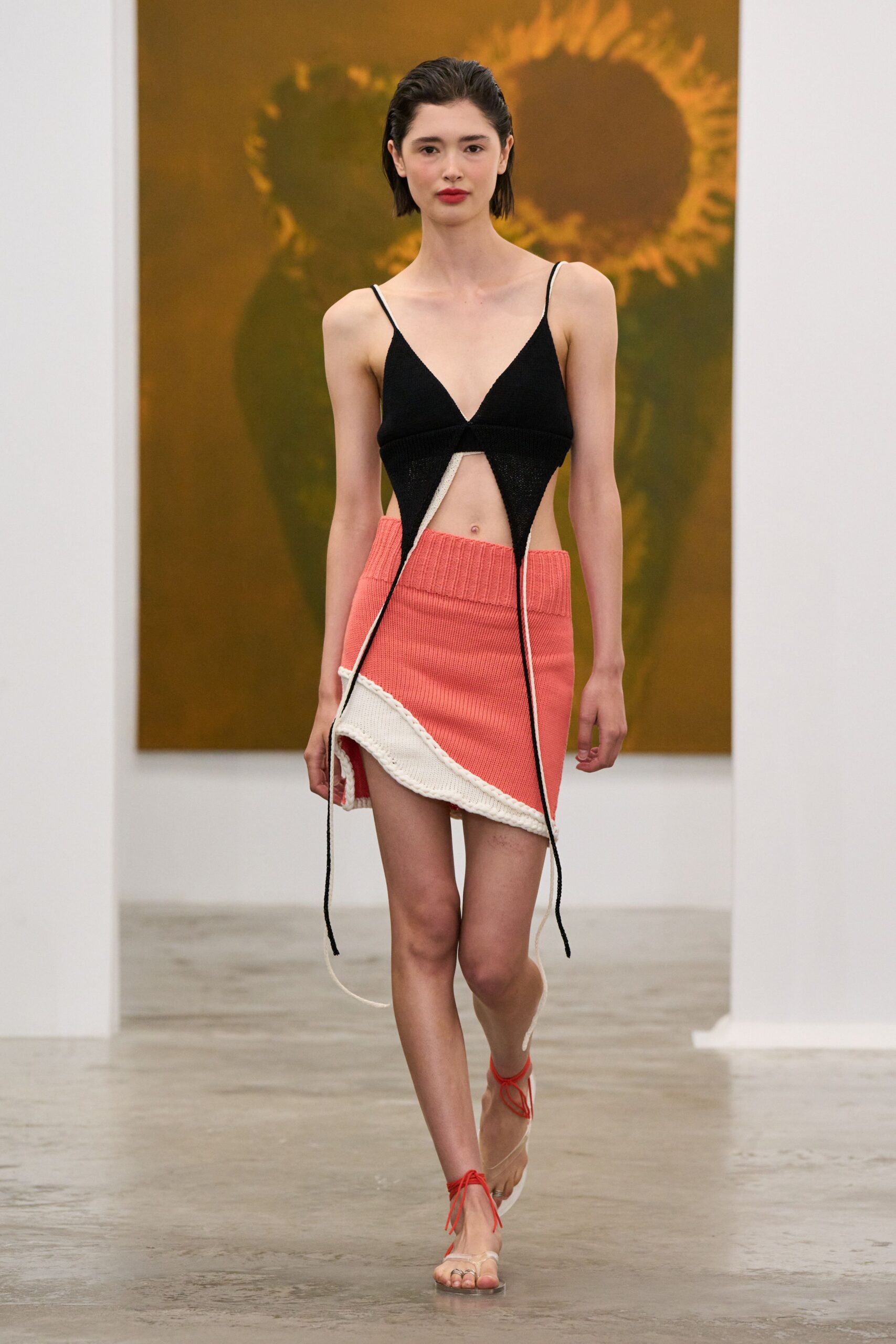 Black ribbed knit bralette top with long contrast cords hanging past the hem. Coral rib-knit mini with a folded waistband and curled cream edge. Clear thong sandals tied at the ankle with red cord. Walking through a museum atrium after the gift shop has closed.